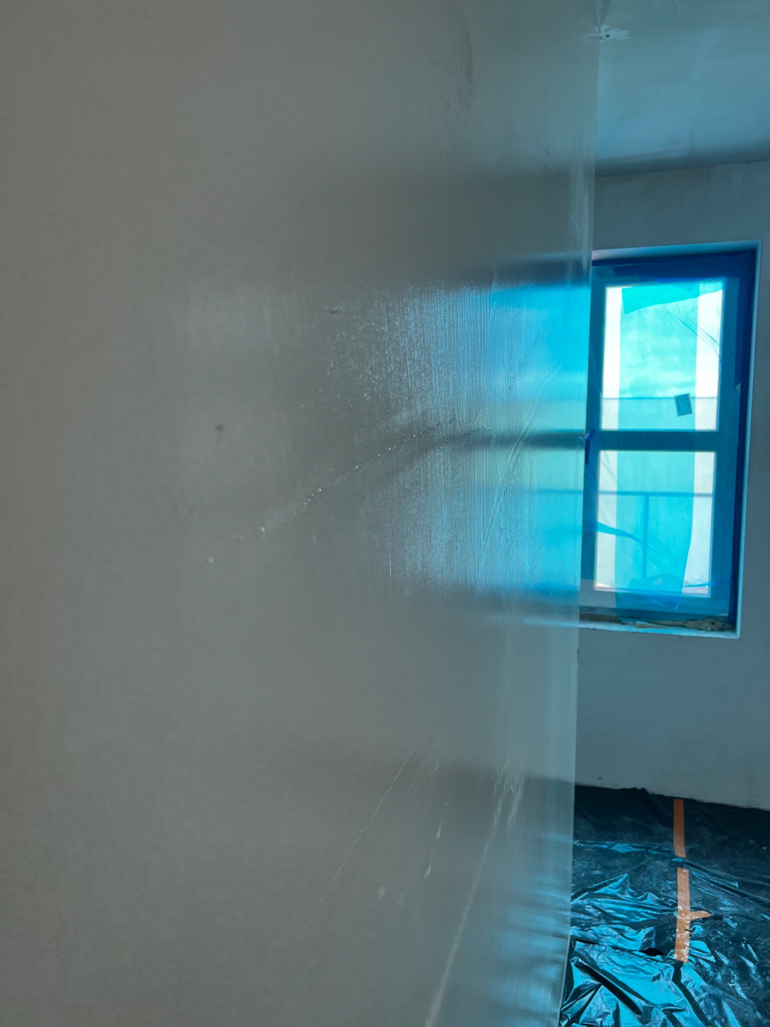 Świeżo nałożona, mokra warstwa na ścianie w pokoju, okno zabezpieczone folią, podłoga przykryta folią ochronną.
