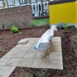 Świeżo posadzone rośliny w ogrodzie z korą, widoczna ławka ogrodowa przykryta folią ochronną, płytki chodnikowe prowadzą wzdłuż rabaty, żółta ściana budynku w tle.