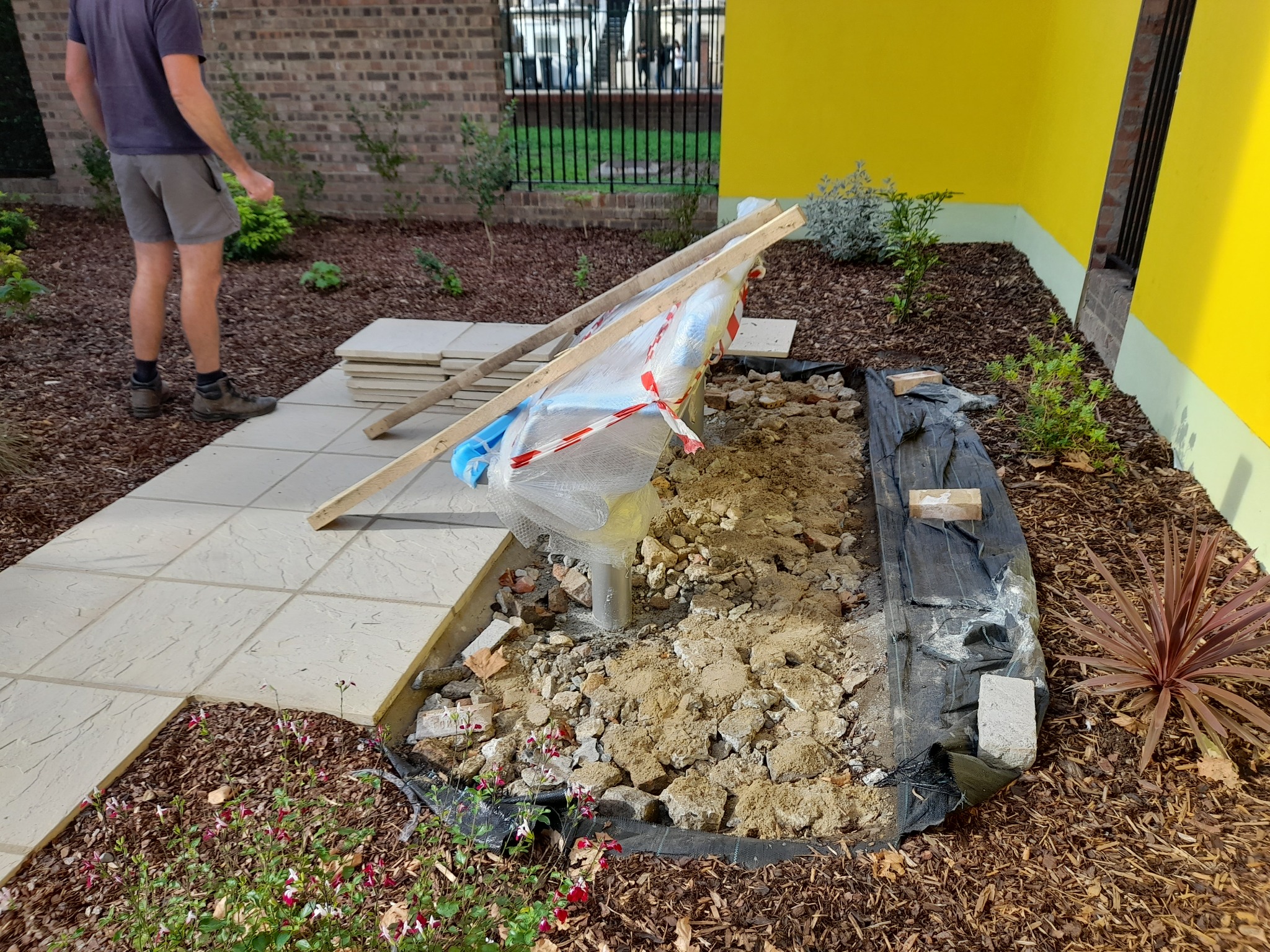 Przygotowanie podłoża pod system nawadniania w ogrodzie: wykop z gruzem, czarną folią, obok ułożone płyty chodnikowe i drewniane stemple, w tle żółta ściana budynku i ogrodzenie.