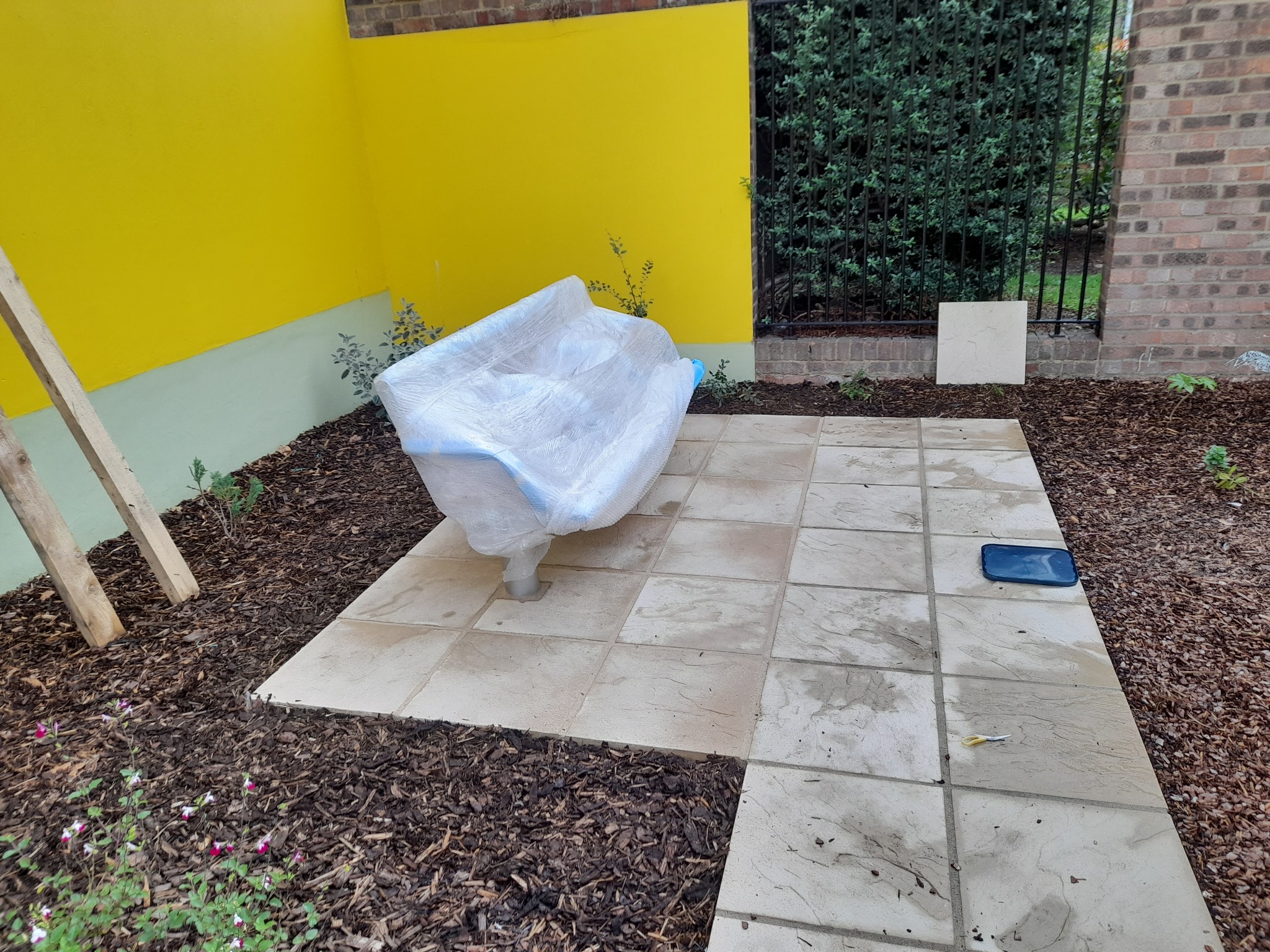 Nowo ułożona, kwadratowa nawierzchnia z jasnych płyt w ogrodzie, z przykrytą folią ochronną fontanną lub rzeźbą pośrodku, obok żółtej ściany i drewnianej podpory, z widocznymi narzędziami...