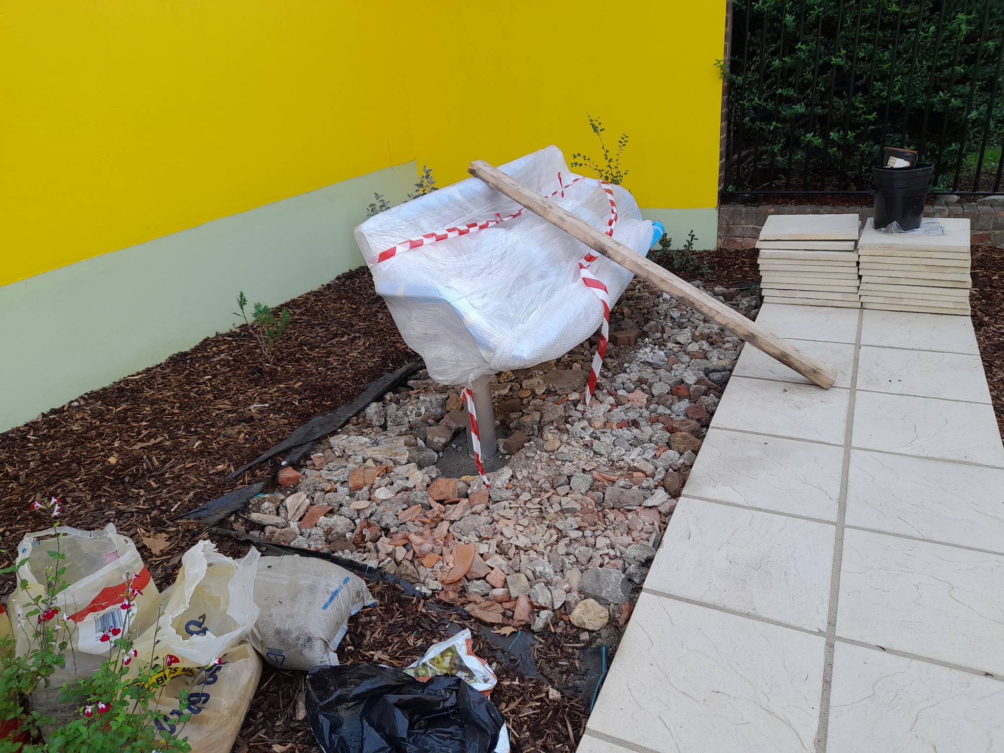 Przygotowanie podłoża pod system nawadniania: wykop z gruzem, rura PCV owinięta taśmą ostrzegawczą, obok ułożone płyty chodnikowe i worki z ziemią, żółta ściana budynku w tle.