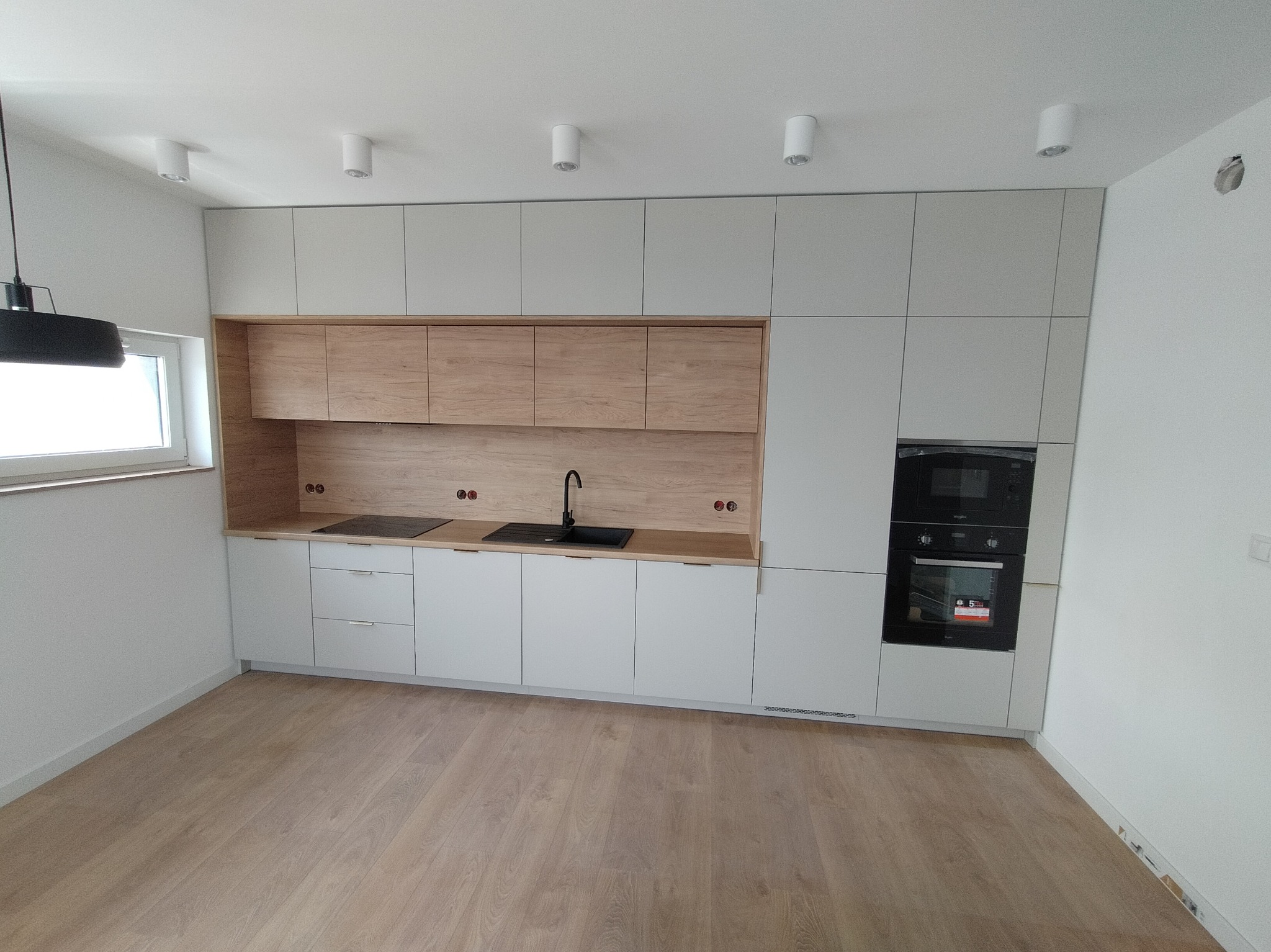 Nowoczesna kuchnia z zabudową w odcieniach bieli i drewna, czarny zlew i płyta grzewcza, jasna podłoga, minimalistyczne oświetlenie sufitowe.