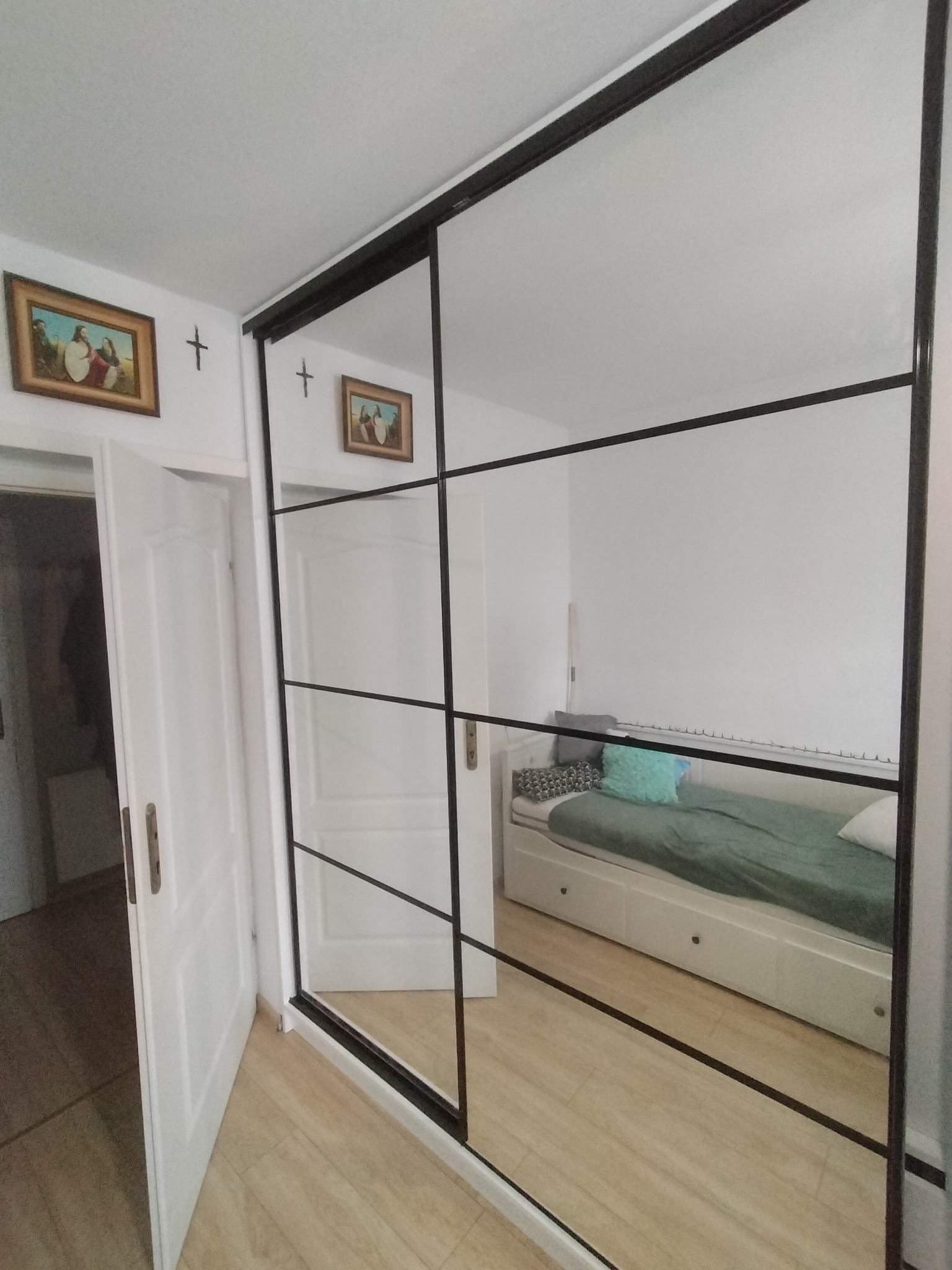 Nowoczesna szafa wnękowa z przesuwnymi drzwiami lustrzanymi z czarnymi szprosami, odbijająca wnętrze pokoju z łóżkiem i otwartymi drzwiami.