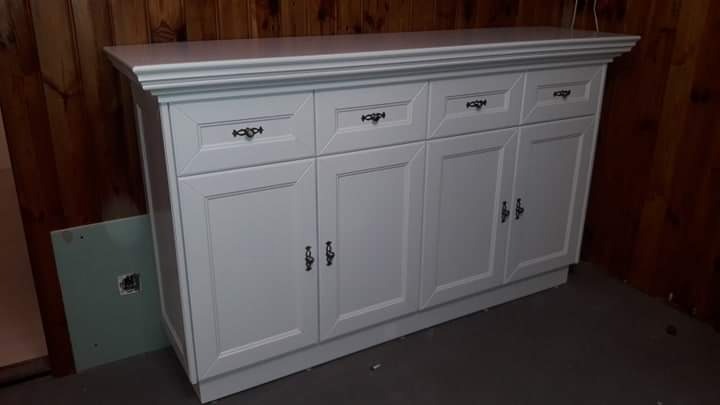 Biała komoda z szufladami i szafkami, z ciemnymi uchwytami, na tle drewnianej ściany, z niedokończonym fragmentem ściany obok.