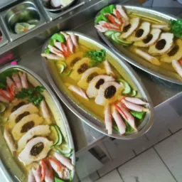 Trzy owalne półmiski z galaretą, plastrami mięsa z ciemnym farszem, plasterkami ogórka, szynką zawiniętą w ruloniki i natką pietruszki, ustawione na metalowym stole, widoczne pojemniki z dodatkami.