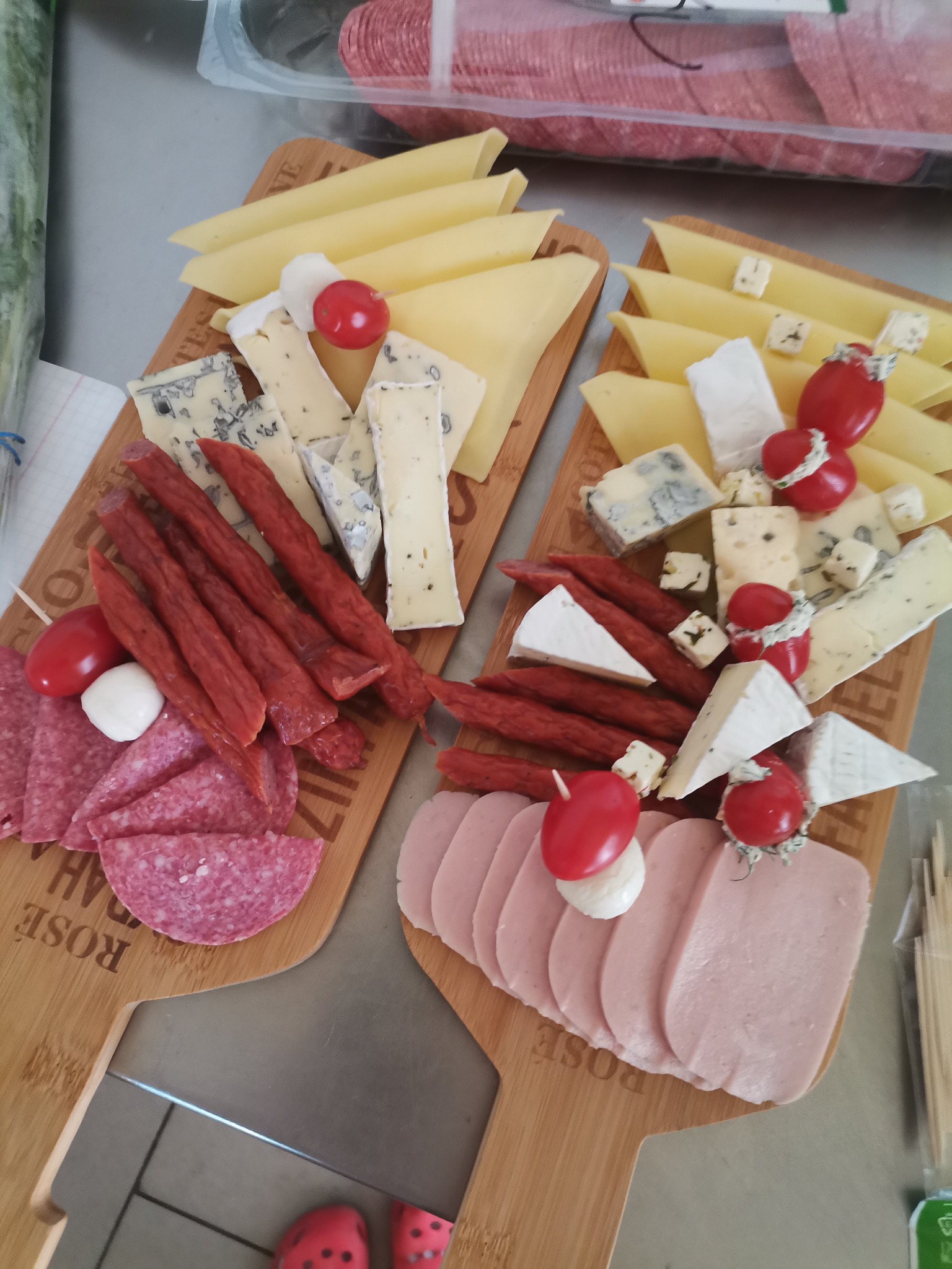 Dwie drewniane deski do serwowania z różnymi rodzajami serów, wędlin, salami, kabanosów i pomidorków cherry z mini mozzarella, ułożone na metalowym blacie.