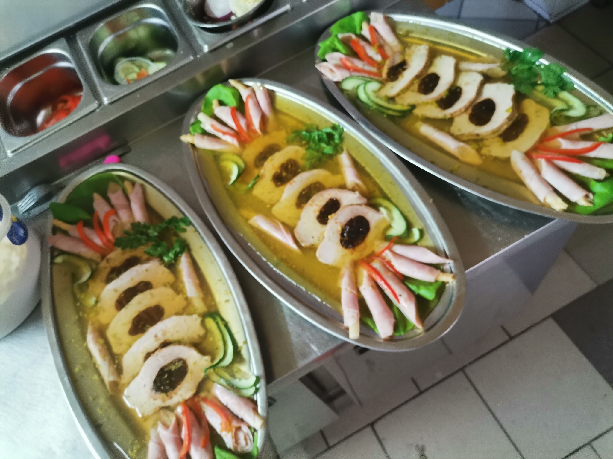 Trzy owalne półmiski z galaretą, plastrami mięsa z ciemnym farszem, plasterkami ogórka, szynką zawiniętą w ruloniki i natką pietruszki, ustawione na metalowym stole, widoczne pojemniki z dodatkami.