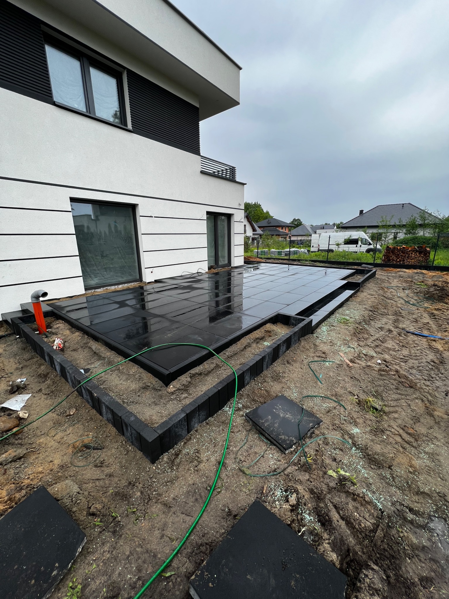 Nowoczesny taras z czarnej kostki w Łodzi, w trakcie układania, z widocznymi obrzeżami i przygotowanym podłożem, na tle białej elewacji budynku.