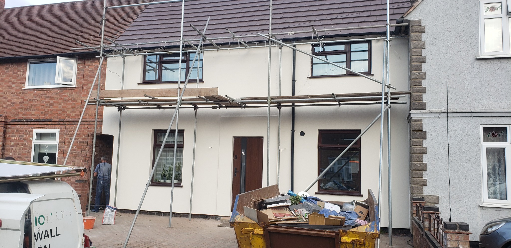 Dom jednorodzinny w trakcie prac elewacyjnych: świeżo otynkowana, kremowa ściana frontowa z brązowymi oknami i drzwiami, otoczona rusztowaniem. Po prawej stronie widoczna elewacja sąsiedniego domu...