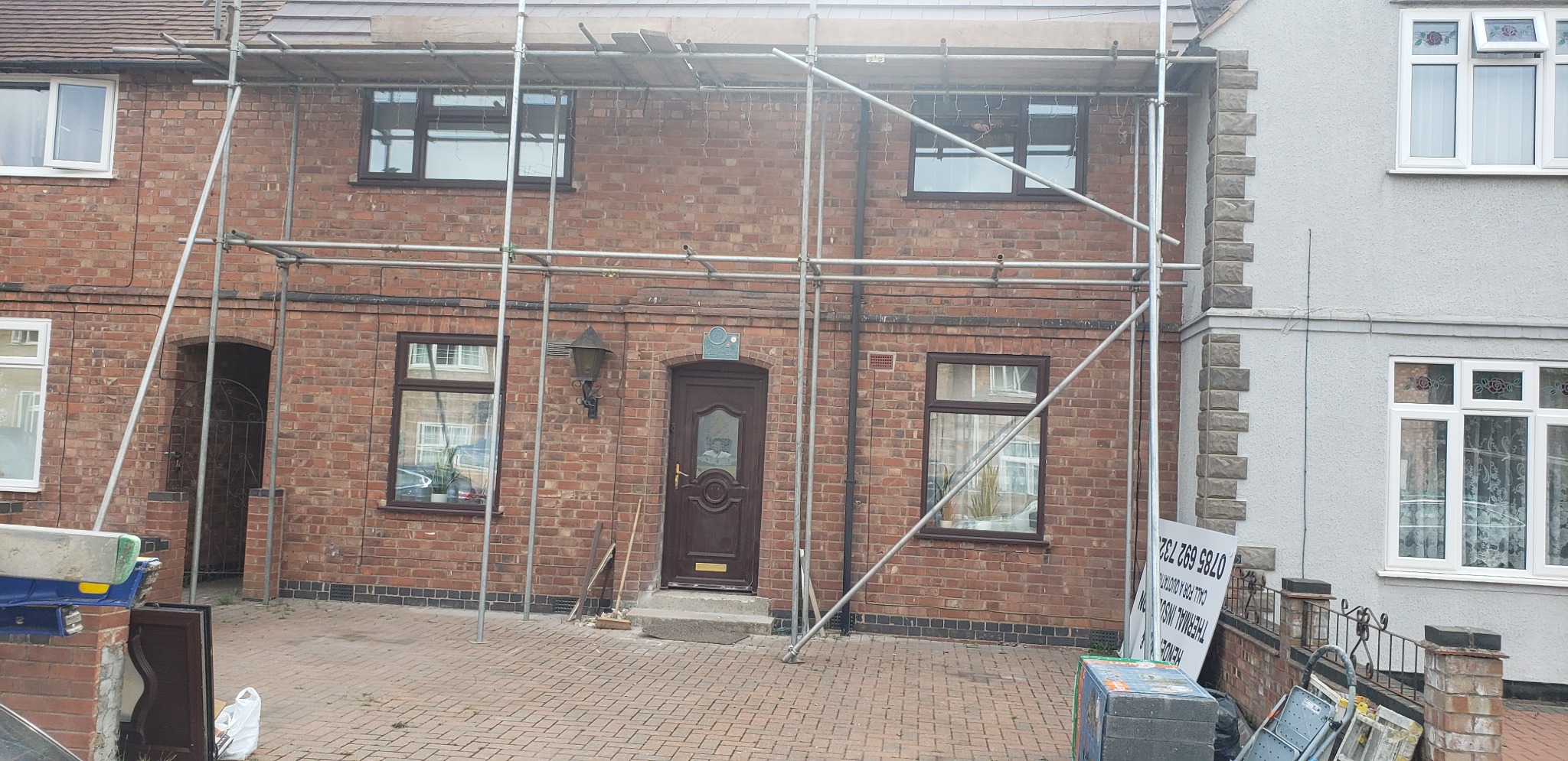 Dom z czerwonej cegły w trakcie renowacji elewacji, otoczony rusztowaniem; widoczny fragment sąsiedniego domu z szarą elewacją i dekoracyjnymi elementami.