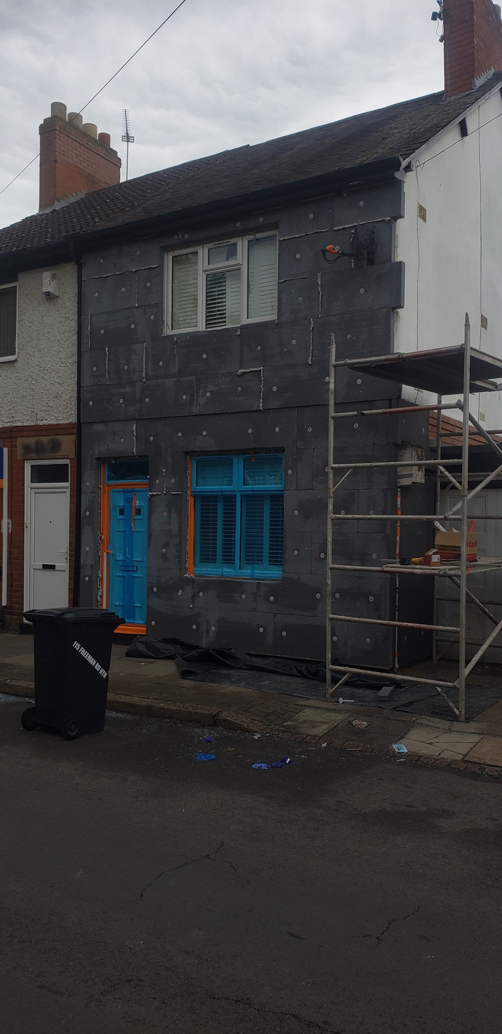 Dom jednorodzinny w trakcie prac izolacyjnych elewacji, widoczne szare płyty izolacyjne, rusztowanie i zabezpieczone okna oraz drzwi folią ochronną, obok stoi czarny kontener na śmieci.