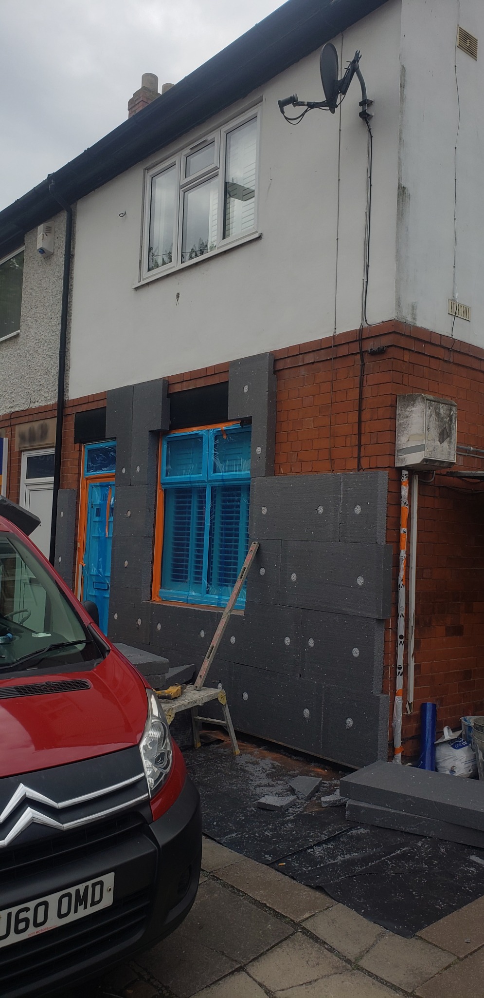 Dom w trakcie nakładania szarej izolacji na elewację, okna zabezpieczone folią, widoczny fragment czerwonego samochodu dostawczego i drabina.