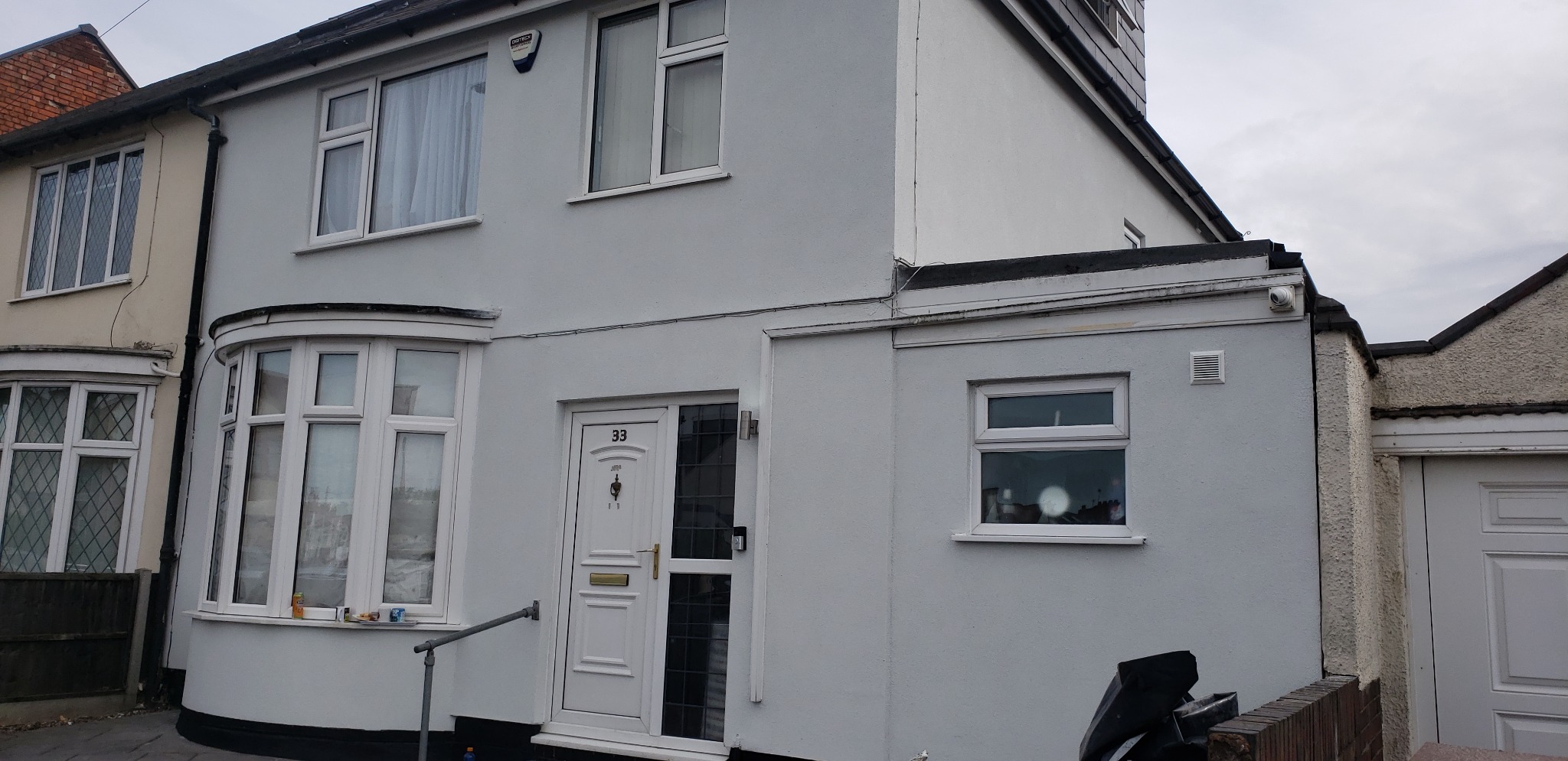Jasnoszary dom jednorodzinny z białymi oknami i drzwiami, widoczny numer domu '33', zmodernizowana elewacja, obok sąsiedni dom z ceglanym dachem.