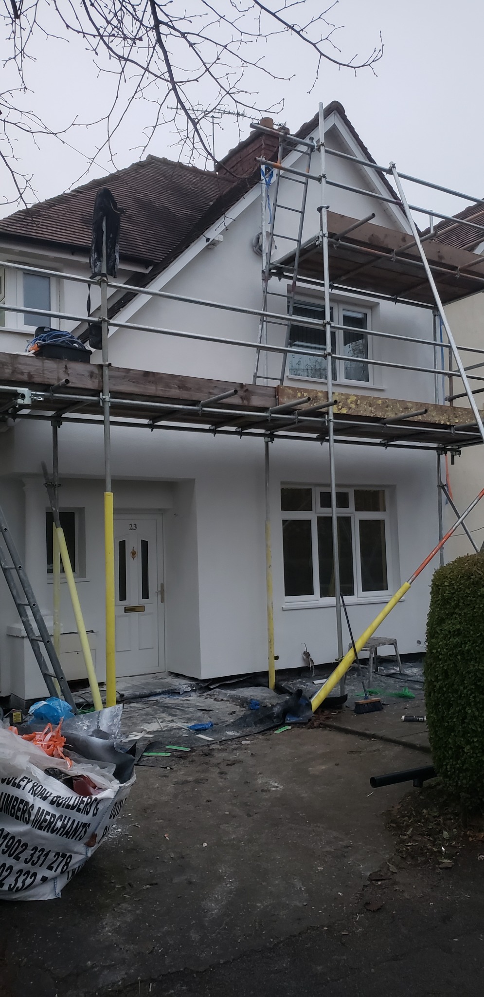 Dom jednorodzinny z białymi ścianami elewacji, w trakcie prac wykończeniowych, widoczne rusztowania i materiały budowlane, numer domu 23 nad drzwiami.
