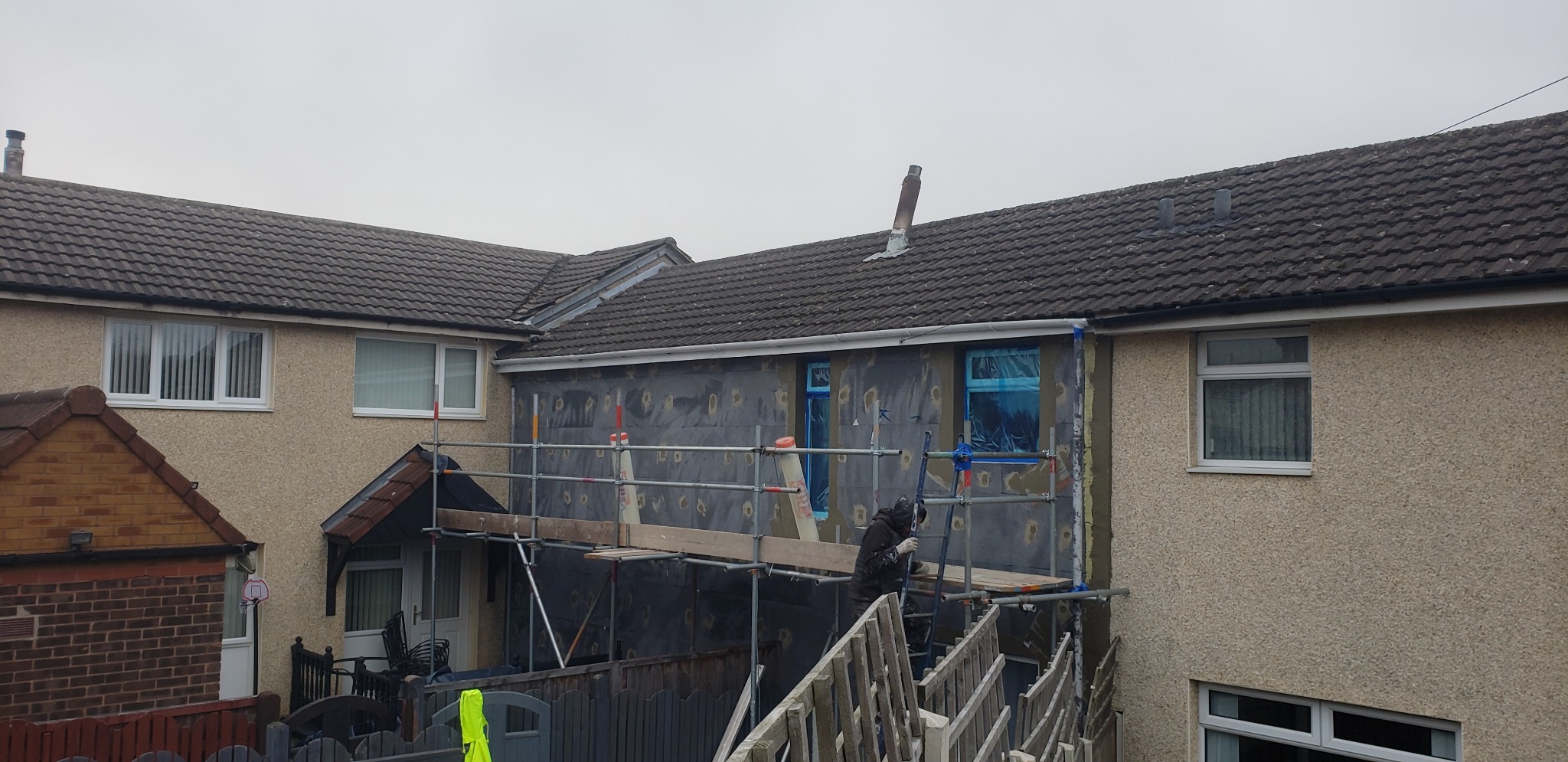 Prace na rusztowaniu przy ocieplaniu elewacji domu jednorodzinnego, okna zabezpieczone folią, widoczne płyty izolacyjne i fragment otynkowanej ściany.