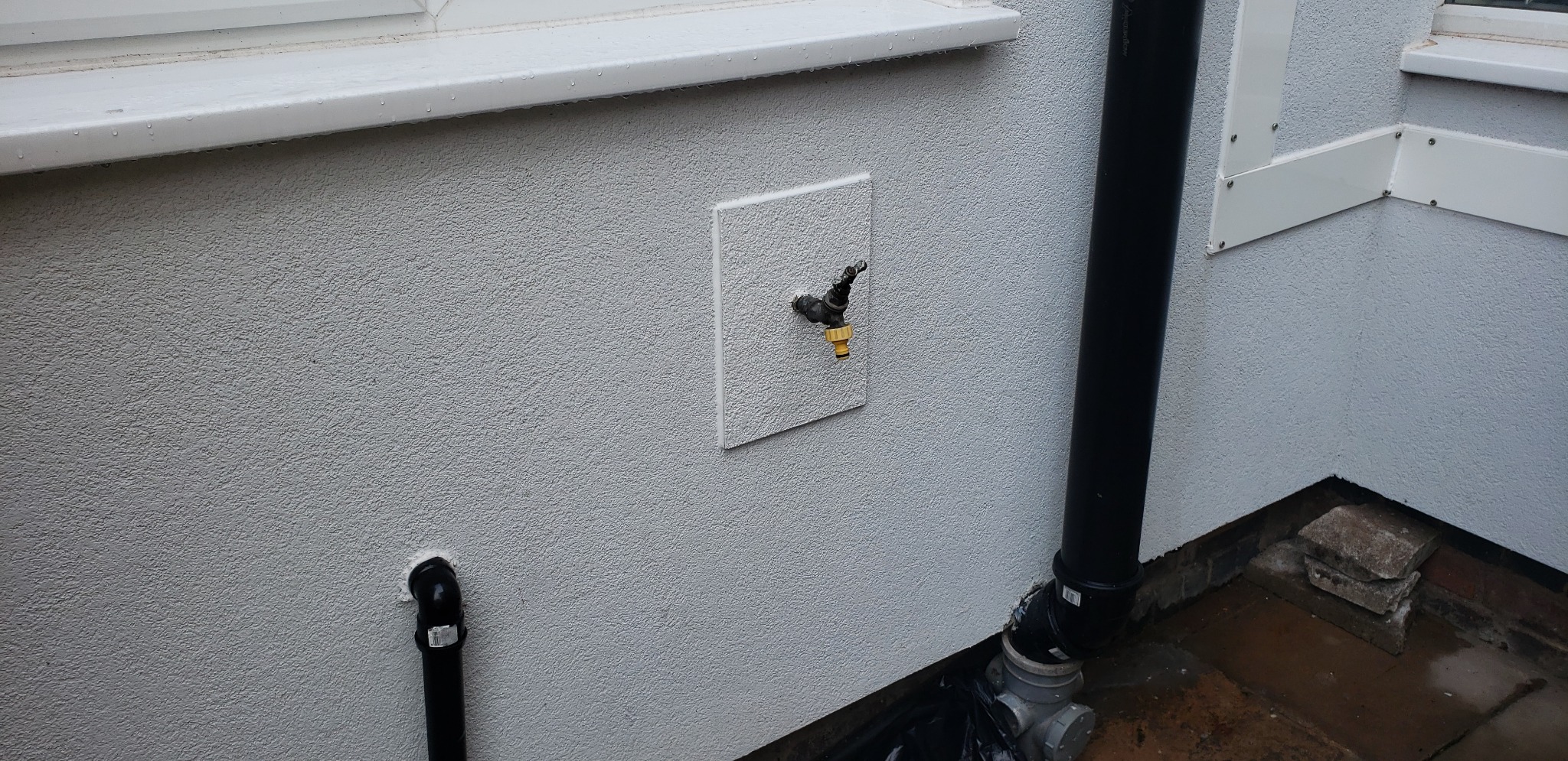 Elewacja budynku po tynkowaniu z widocznym kranem na białej płytce i czarną rurą spustową, wykończona pod oknem i przy ziemi.