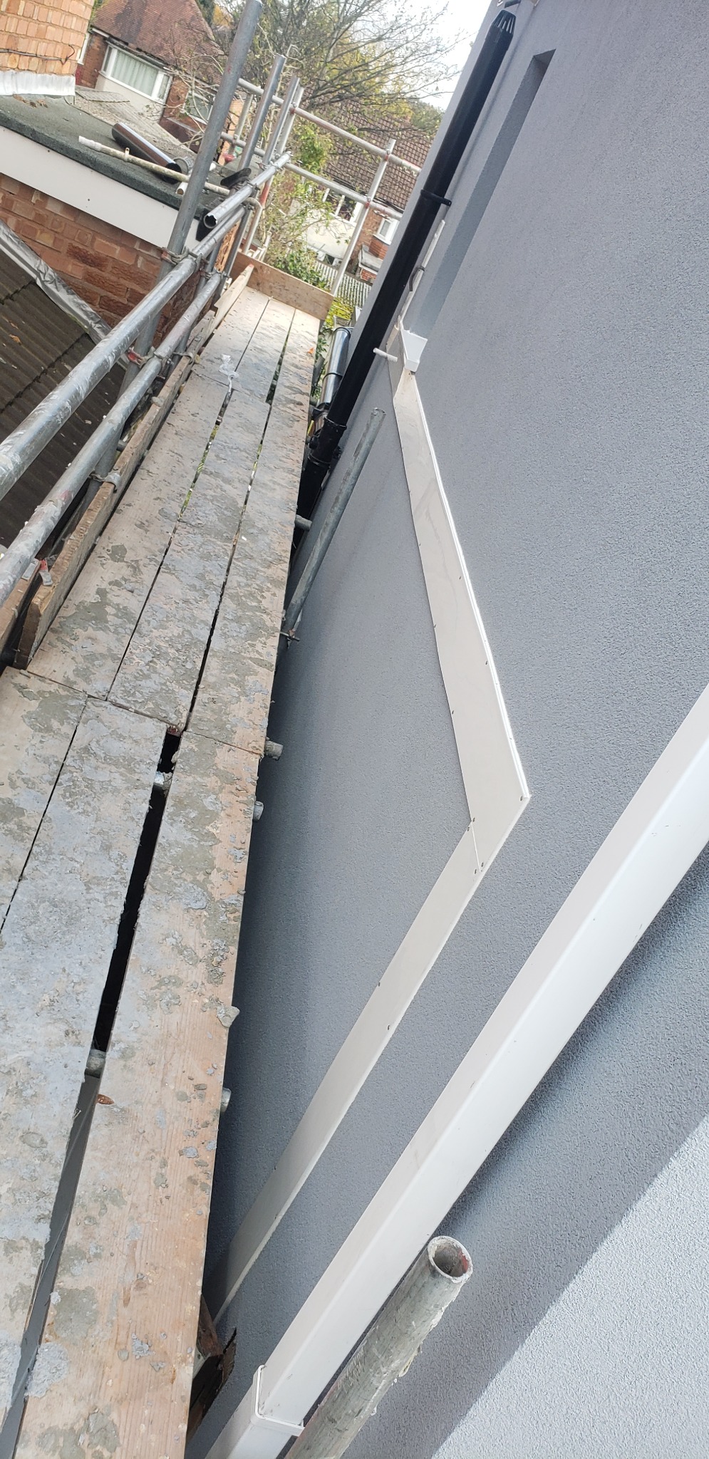 Szarego koloru elewacja budynku z zamontowanymi białymi elementami wykończeniowymi i rurą spustową, widok z rusztowania.