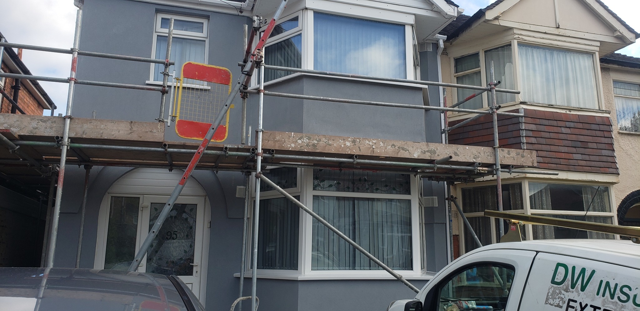 Szare, odnowione ściany domu w zabudowie szeregowej, widoczne rusztowanie z pomarańczowym zabezpieczeniem, sąsiedni dom z brązową okładziną pod oknem.