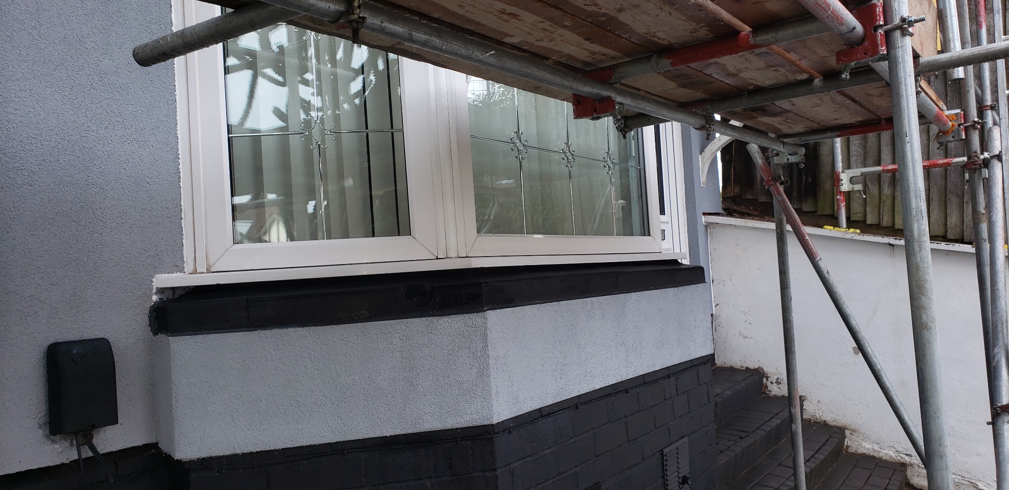 Fragment elewacji budynku z szarym tynkiem strukturalnym, białą ramą okienną z dekoracyjnymi szybami i czarnym cokołem, widoczne rusztowanie.
