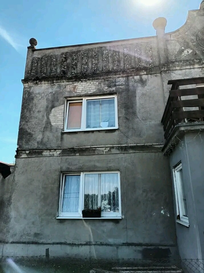 Dwupiętrowy budynek z szarą elewacją wymagającą odświeżenia, widoczne okna z firankami i balkon z drewnianą balustradą.