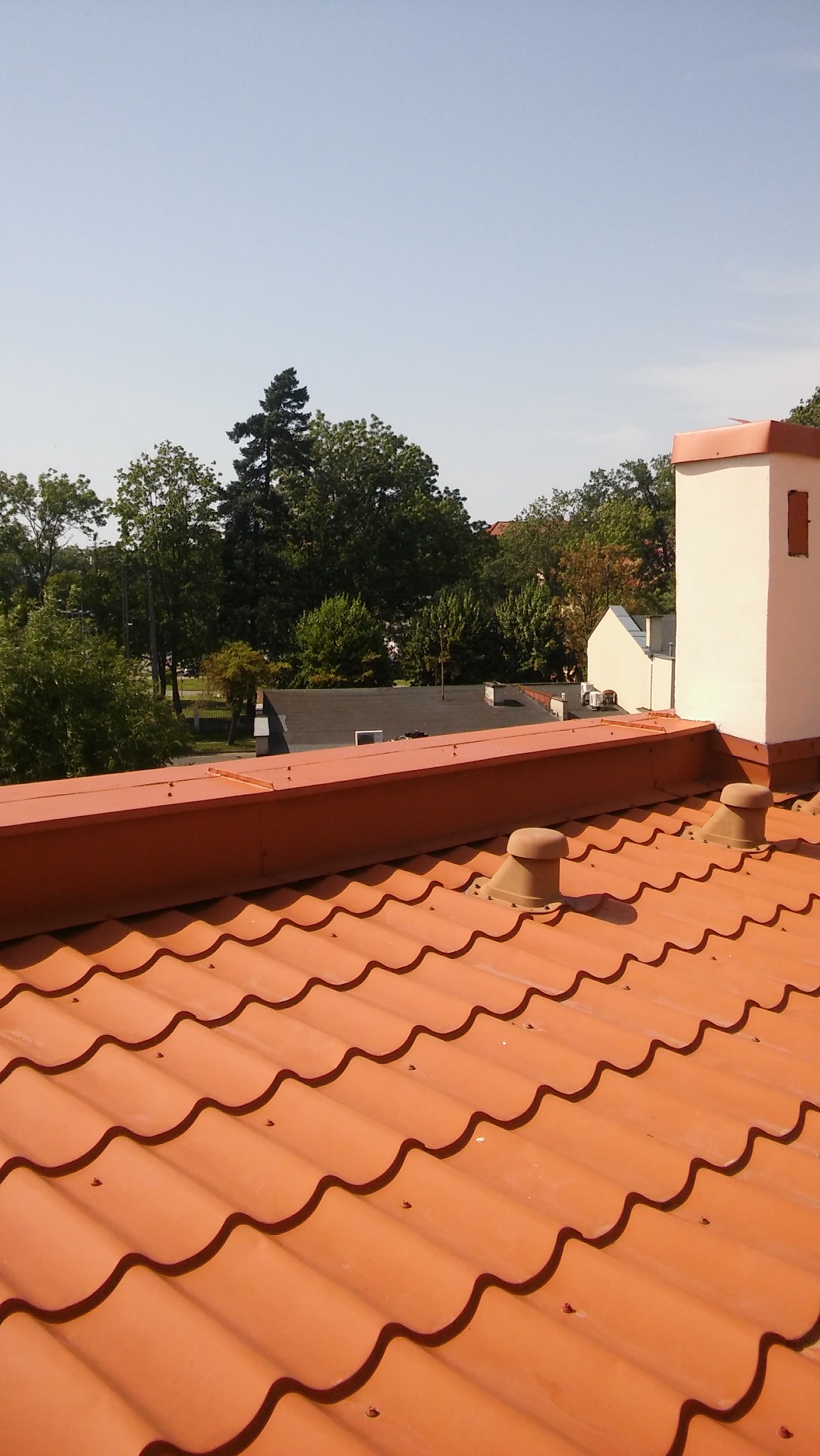 Pomarańczowa dachówka ceramiczna z kominem w tle, widok z góry na tle zieleni i błękitnego nieba.