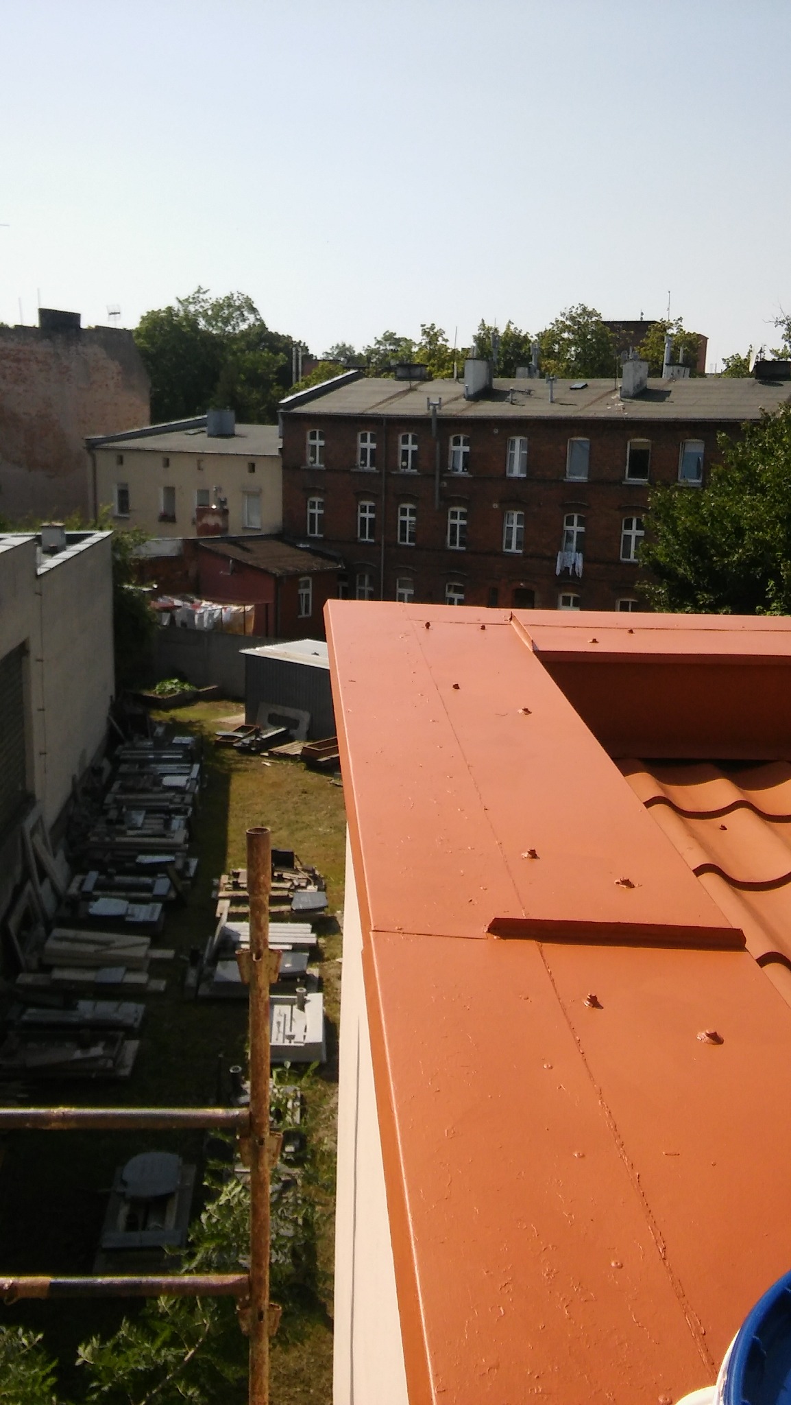 Świeżo pomalowana, pomarańczowa obróbka blacharska dachu z widocznymi nitami, na tle budynku mieszkalnego i zieleni.