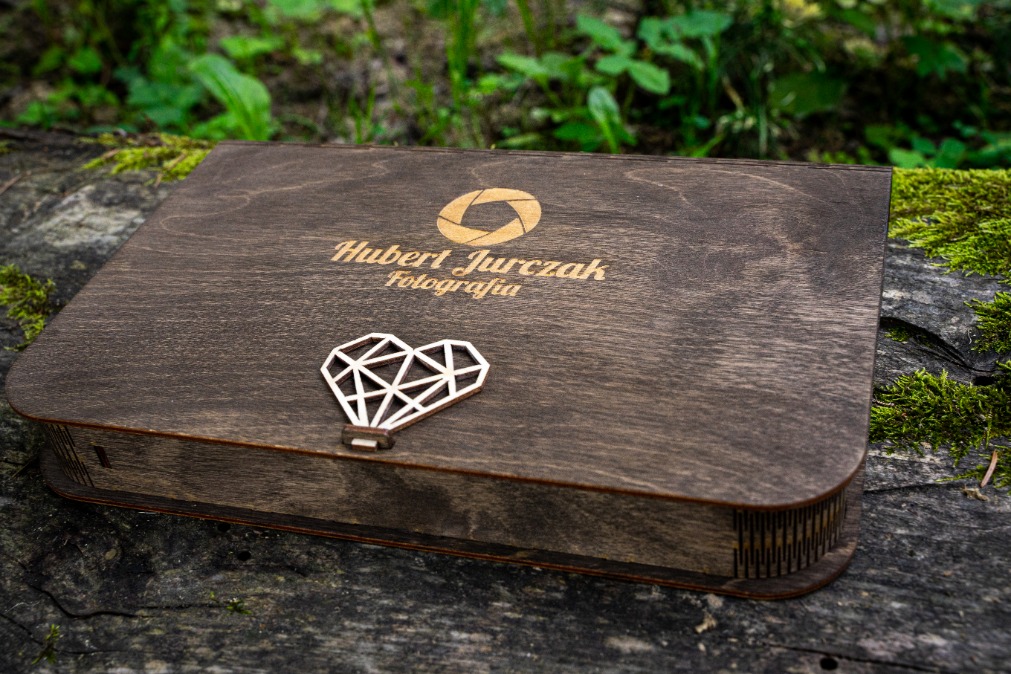 Ciemnobrązowe, drewniane pudełko z wygrawerowanym logo fotografa 'Hubert Jurczak Fotografia' i ozdobnym, geometrycznym zamknięciem w kształcie serca-balonika, leżące na zewnątrz na zarośniętym...