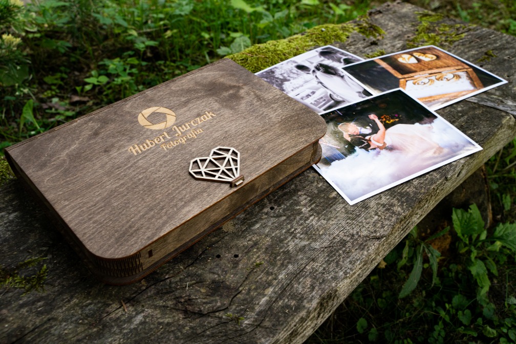 Drewniane pudełko z logo fotografa 'Hubert Jurczak Fotografia' i geometrycznym sercem jako zapięciem, leżące obok wydrukowanych zdjęć na spróchniałej ławce w plenerze.