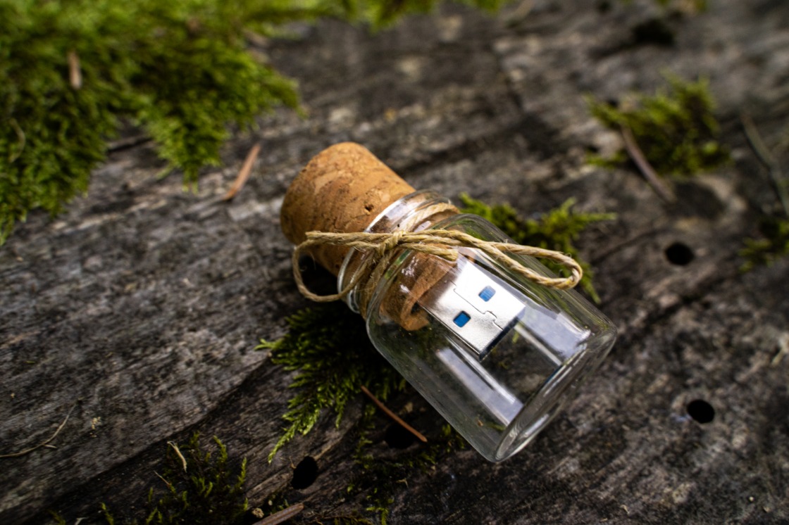 Szklana fiolka z korkiem wypełniona pendrivem USB, przewiązana sznurkiem, leży na szorstkim, ciemnym drewnie pokrytym mchem.