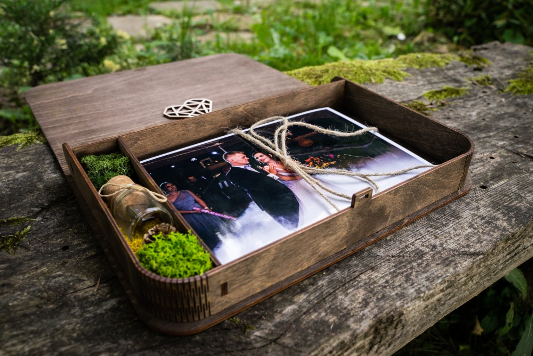 Drewniane pudełko z przegródkami, zawierające zdjęcie pary młodej, małą buteleczkę z korkiem, mech i dekorację w kształcie serca, leżące na drewnianym stole na zewnątrz.
