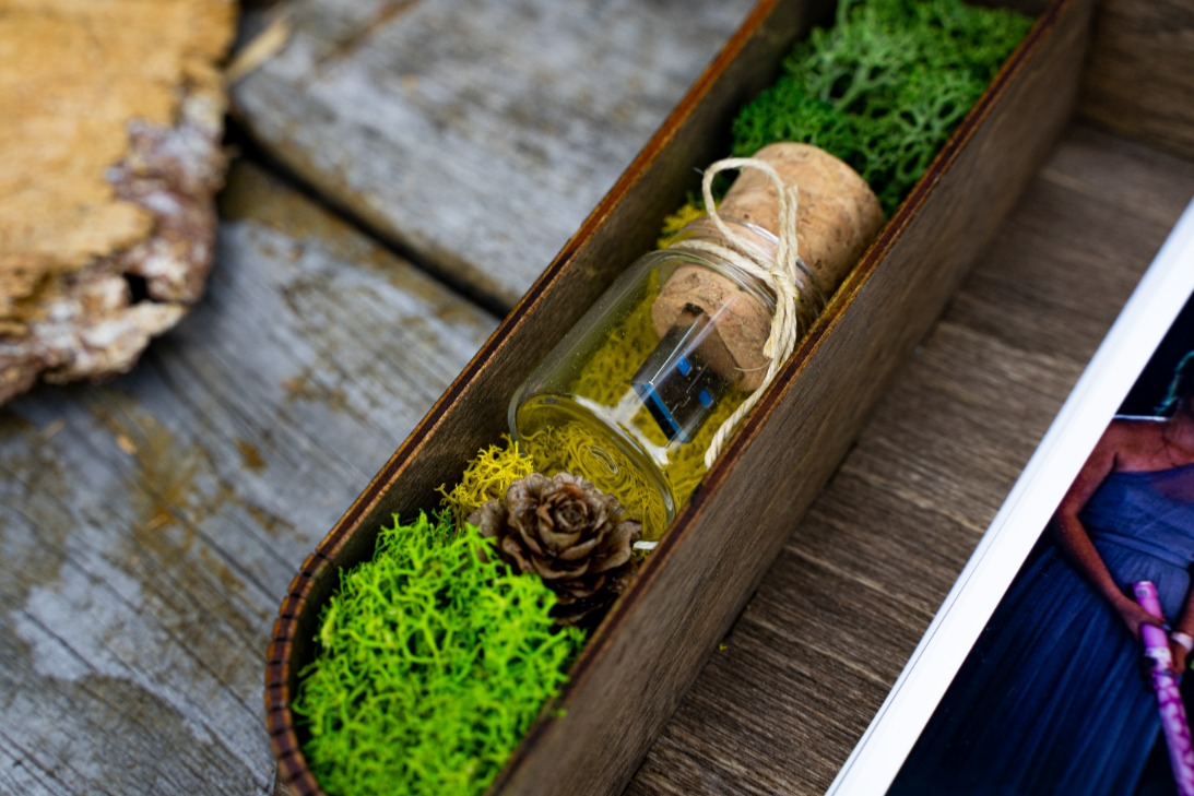 Pamięć USB w szklanej buteleczce z korkiem, ozdobiona sznurkiem, umieszczona w drewnianym pudełku wypełnionym mchem i szyszką, obok widoczne fragmenty zdjęć.