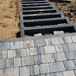 Budowa schodów z elementów betonowych i kostki brukowej na zewnątrz, widoczne niedokończone stopnie i paleta z kostką w tle.