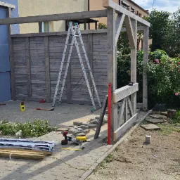 Budowa drewnianej konstrukcji z elementami kostki brukowej w ogrodzie, widoczna drabina, poziomica, wkrętarki i narzędzia brukarskie.