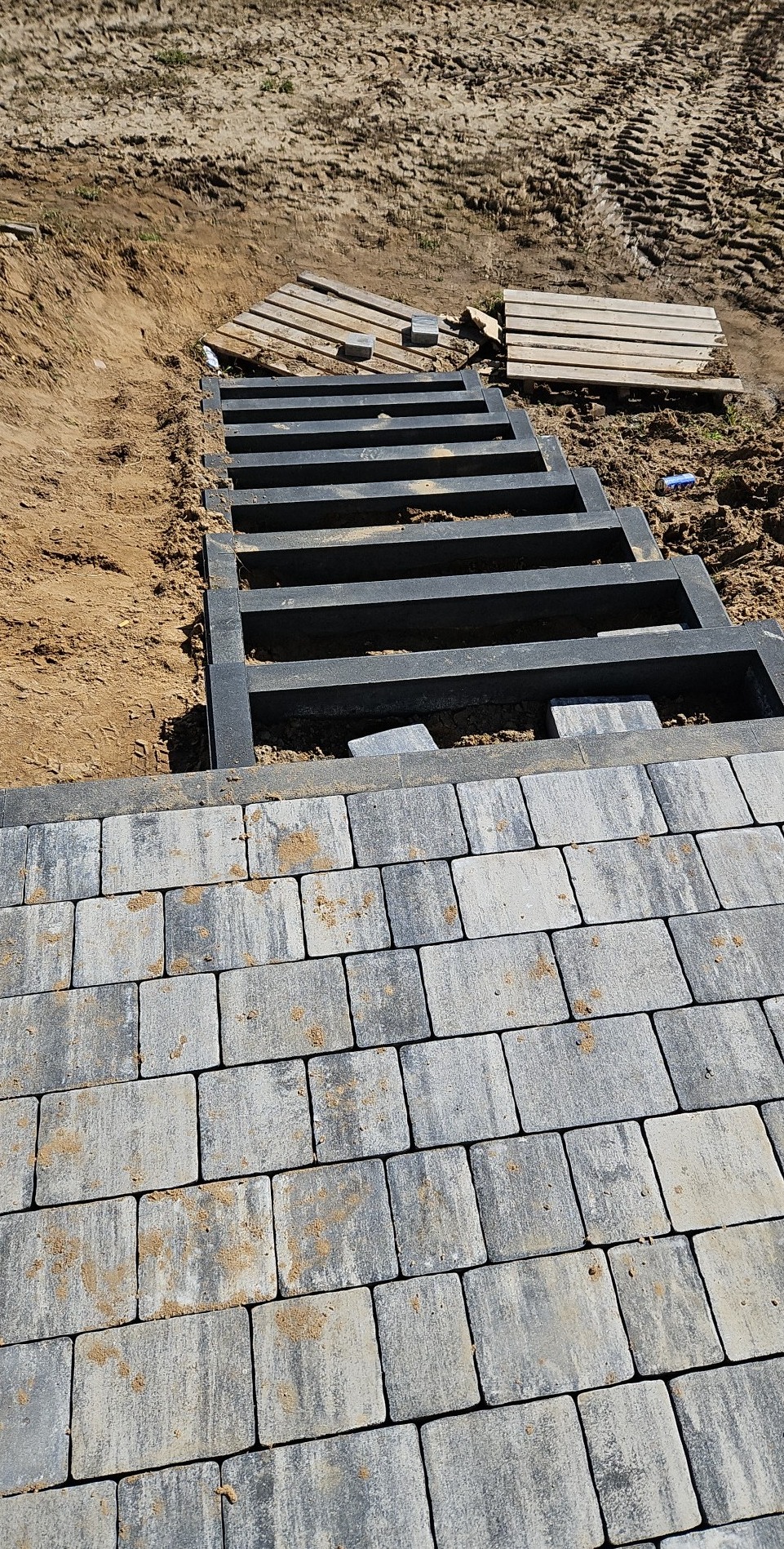 Budowa schodów z elementów betonowych i kostki brukowej na zewnątrz, widoczne niedokończone stopnie i paleta z kostką w tle.