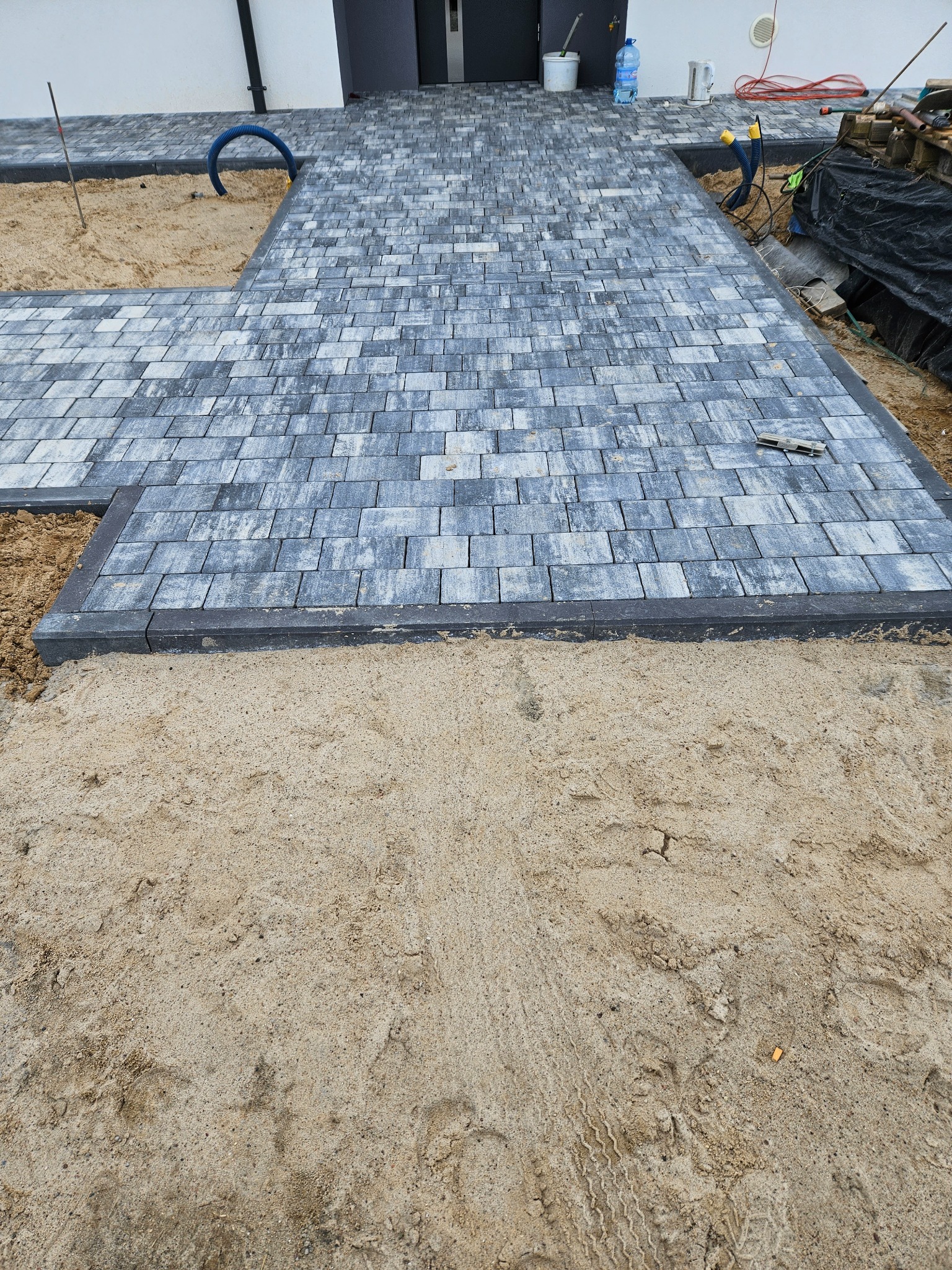 Świeżo ułożona kostka brukowa w odcieniach szarości prowadząca do nowoczesnego domu, widoczny piasek, obrzeża, narzędzia oraz niebieski wąż.