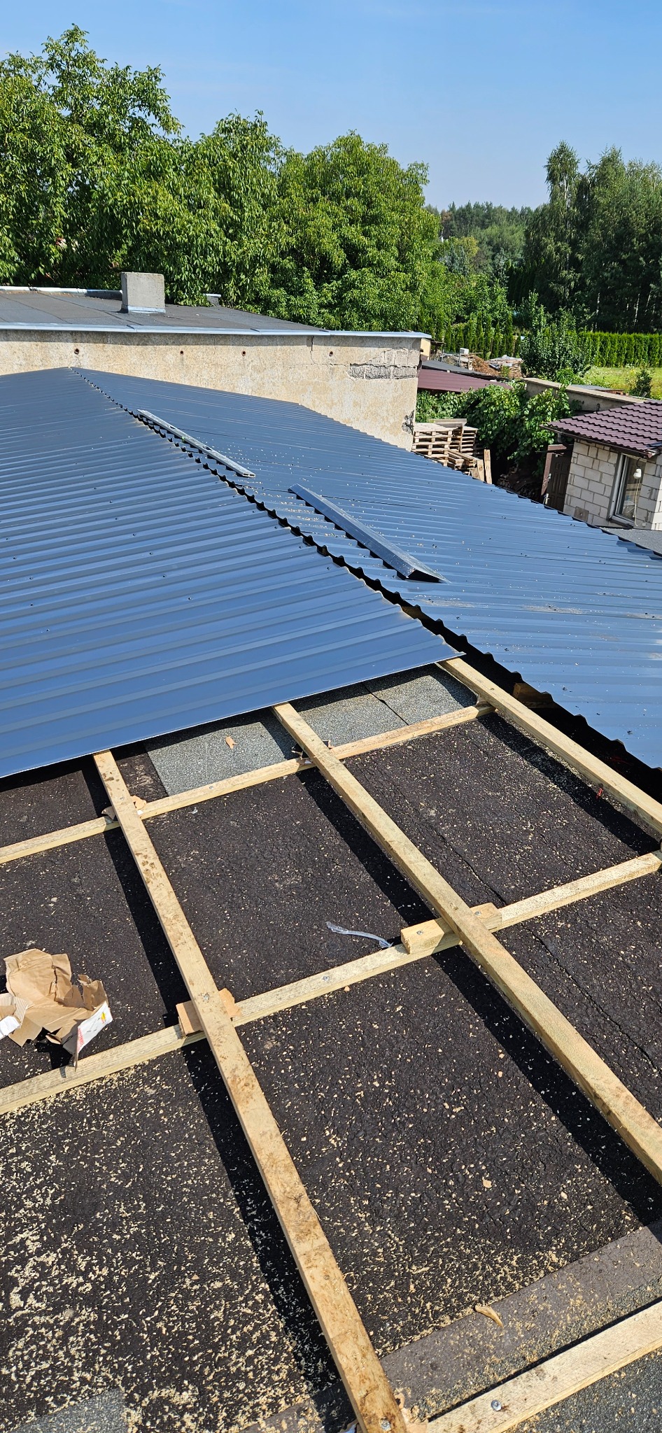 Montaż blachy trapezowej na dachu: widoczne drewniane łaty, fragment pokrycia papą i częściowo ułożona blacha w kolorze grafitowym, w tle zieleń drzew i zabudowania.