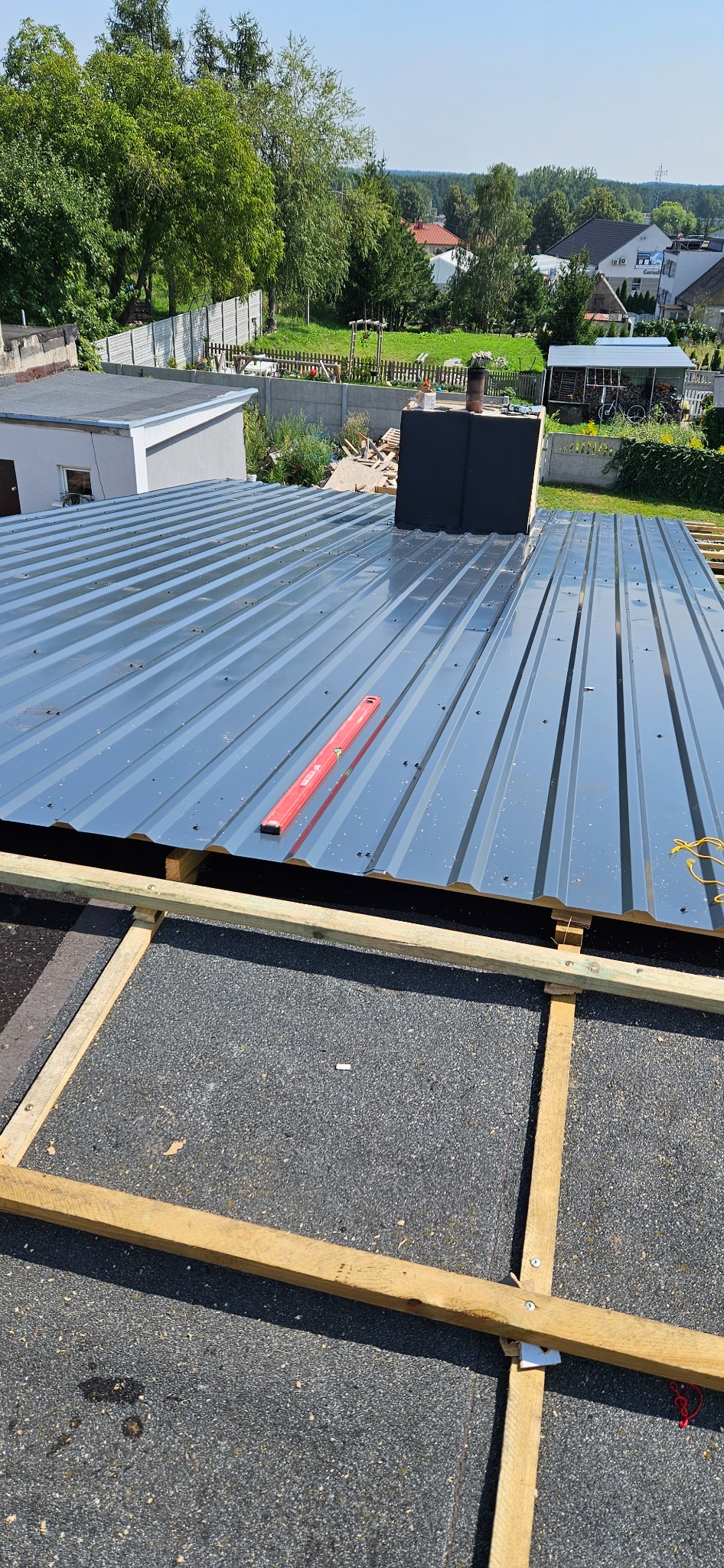 Montaż blachy trapezowej na dachu budynku z kominem, poziomica na dachu, widoczna drewniana konstrukcja szkieletowa.