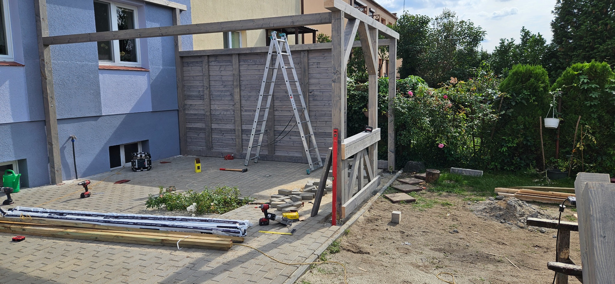 Budowa drewnianej pergoli przy domu z częściowo ułożoną kostką brukową, widoczne narzędzia i materiały budowlane, drabina oparta o drewnianą ścianę.