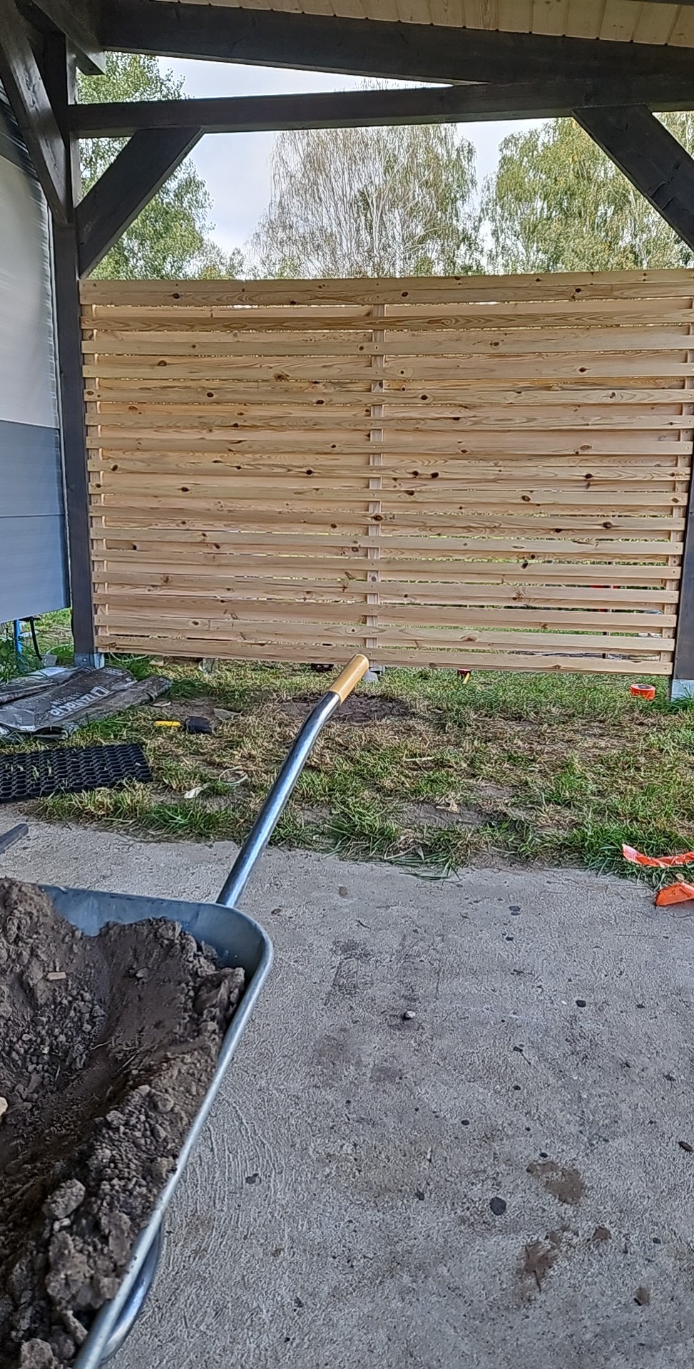 Montaż drewnianego ogrodzenia panelowego z poziomych desek, widoczny wózek z ziemią i narzędzia na trawie.