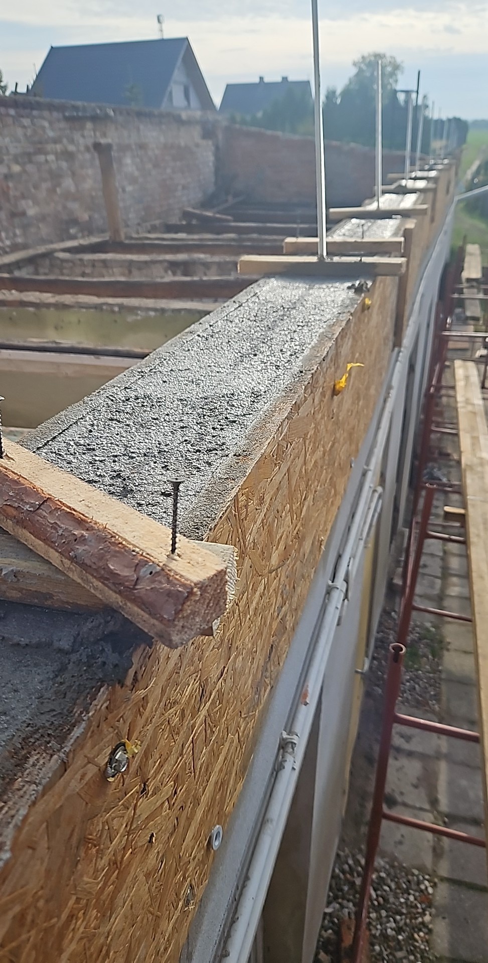 Wylewka betonowa na szalunku z płyty OSB, widoczne gwoździe i fragment drabiny, w tle budynek w trakcie budowy.