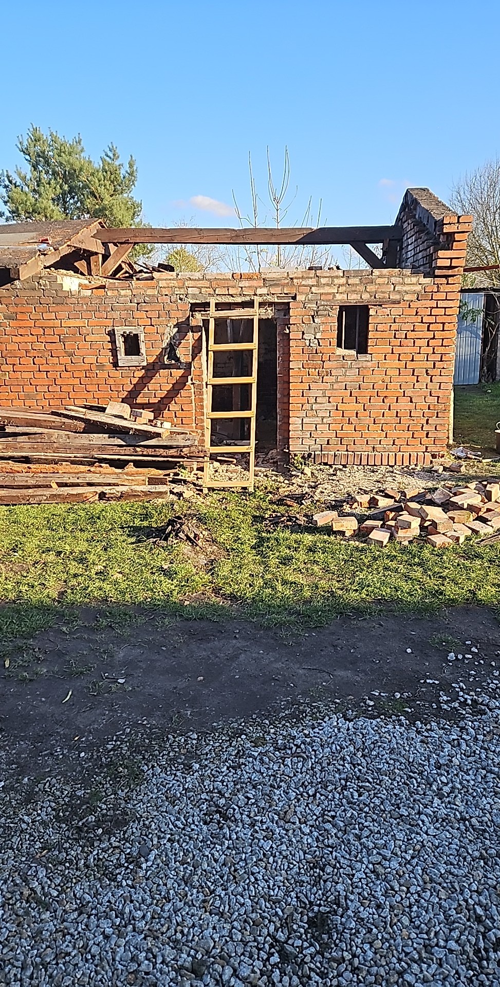 Ceglany budynek w trakcie rozbiórki, widoczne odsłonięte belki stropowe, sterta cegieł i drewniana drabina oparta o wejście, otoczenie zarośnięte trawą, w tle drzewa.