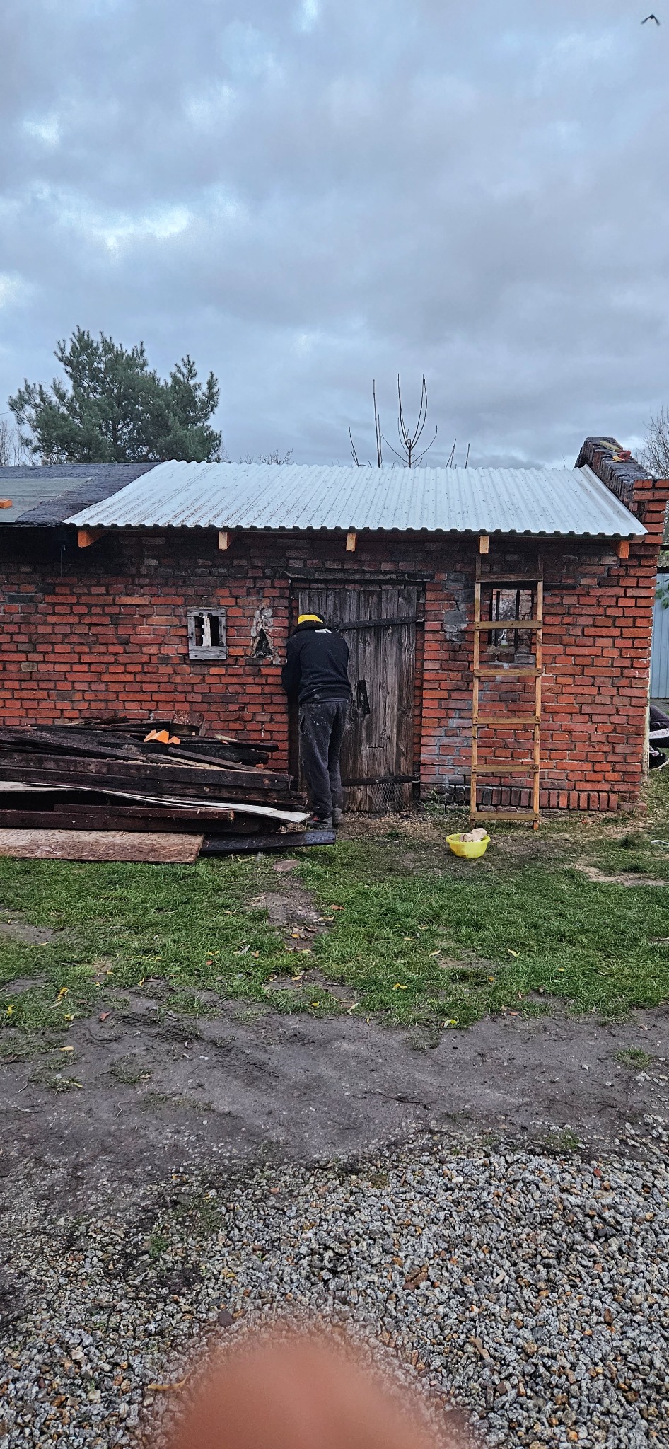 Pracownik w Wągrowcu, w czarnej bluzie i żółtej czapce, muruje ceglaną ścianę budynku gospodarczego z drewnianymi drzwiami i blachą falistą na dachu.
