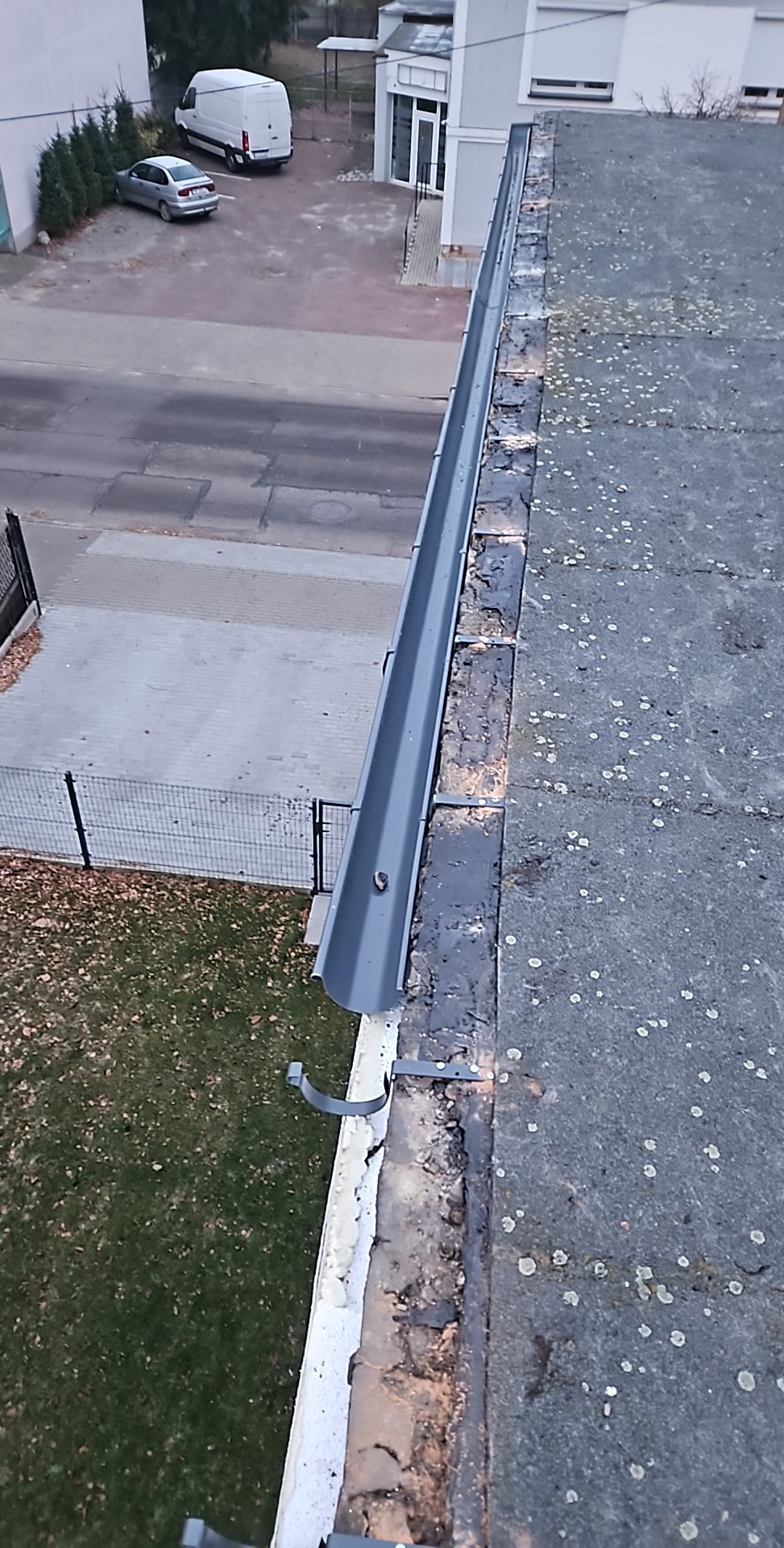 Nowa, szara rynna zamontowana na krawędzi płaskiego dachu, widok z góry na ulicę, samochody i budynki w tle.