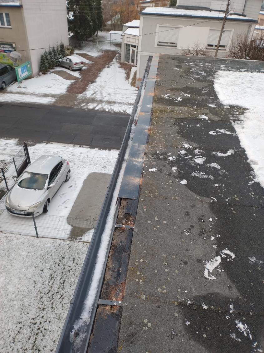Widok z góry na fragment dachu pokrytego papą z widoczną rynną częściowo wypełnioną śniegiem; w tle budynki i zaparkowane samochody w zimowej scenerii.