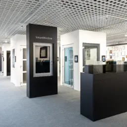 Wnętrze salonu wystawowego z oknami i drzwiami, prezentacja modeli na ekspozytorach, widoczne różne style i materiały wykończeniowe, ściana z certyfikatami w tle.