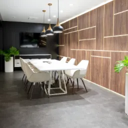 Nowoczesna sala konferencyjna z przesuwnymi szklanymi drzwiami, białym stołem i krzesłami, drewnianą ścianą z podświetleniem LED oraz roślinami w białych donicach.