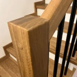 Fragment drewnianych schodów z poręczą z czarnymi, pionowymi elementami i widocznym połączeniem elementów drewnianych na słupku.