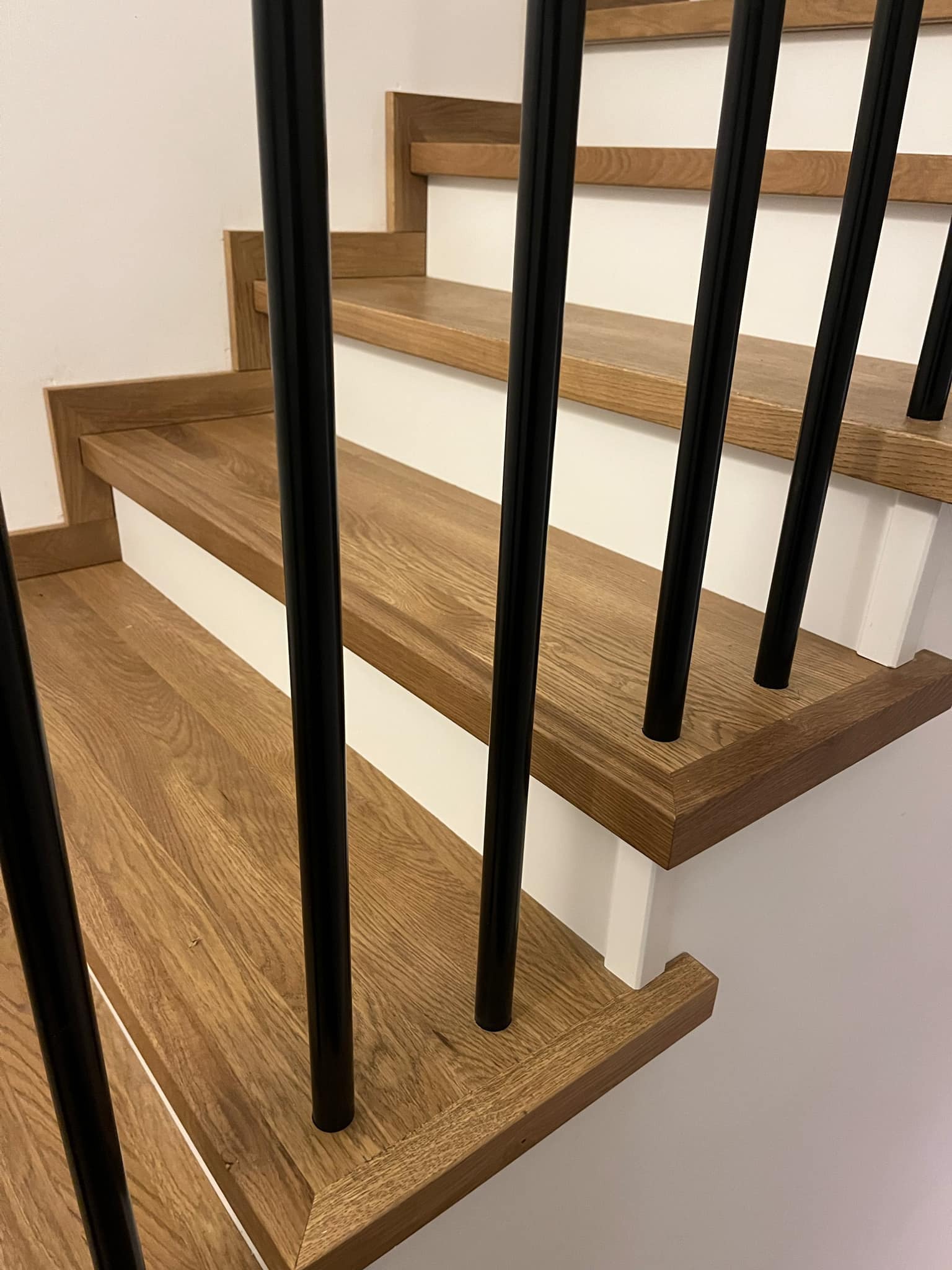Fragment drewnianych schodów z czarnymi, okrągłymi balustradami, widoczne usłojenie drewna i białe podstopnice.