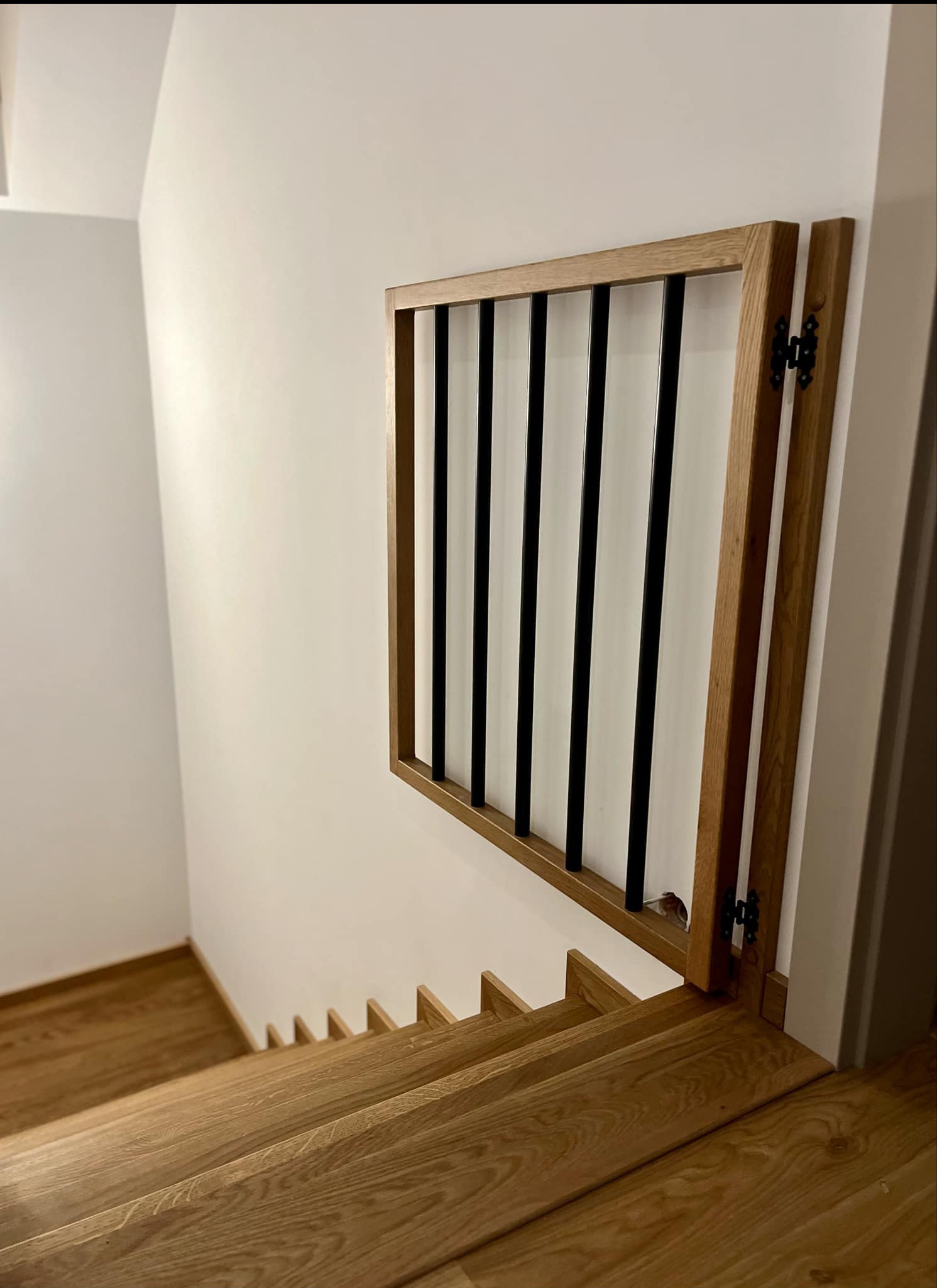 Widok z góry na drewniane schody z zabezpieczeniem w postaci drewnianej ramy z czarnymi, pionowymi prętami, zamontowanej na zawiasach.