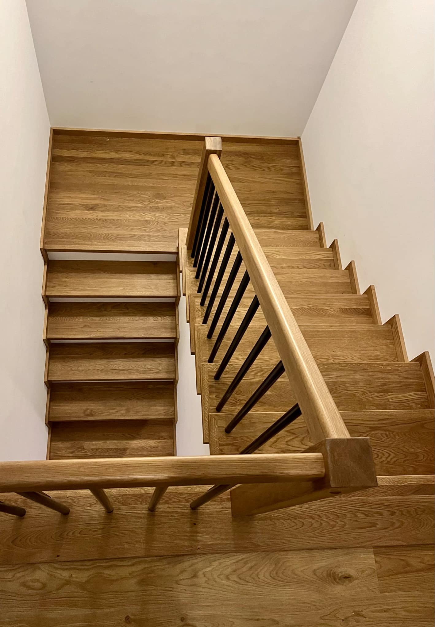 Widok z góry na jasne, dębowe schody z czarnymi, metalowymi balustradami, prowadzące w dół, z wyraźnym usłojeniem drewna i minimalistycznym wykończeniem ścian.